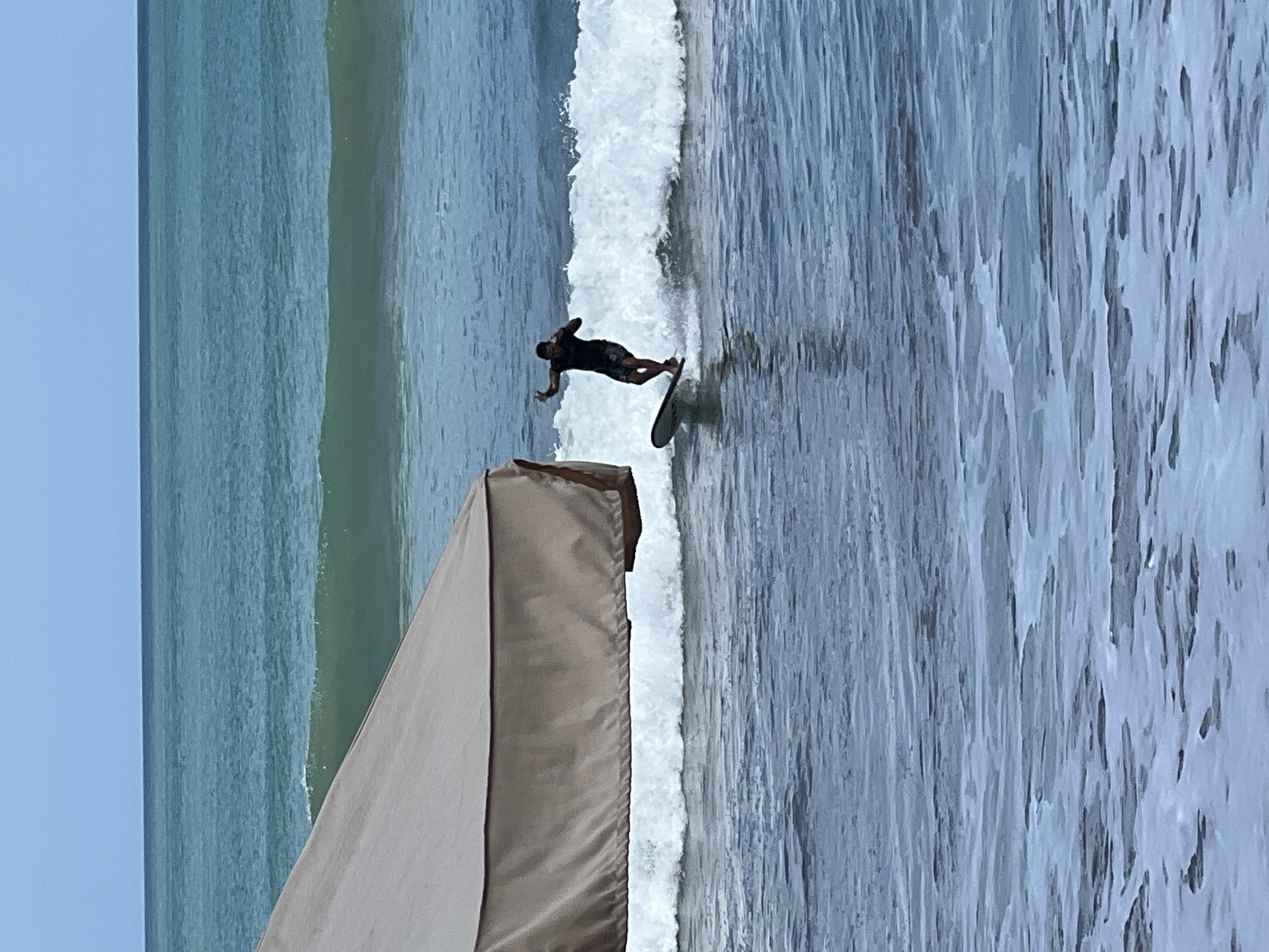 A single surfer, a male wearing a dark shirt and patterned shorts, rides a wave near the shore in Kuta, Indonesia. He is positioned on a surfboard with his arms extended. In the foreground, a large, light-brown tarp, possibly part of a beach umbrella or seating area, partially obscures the left side of the frame. The ocean is choppy with white waves breaking. The sky is overcast with a pale blue hue.