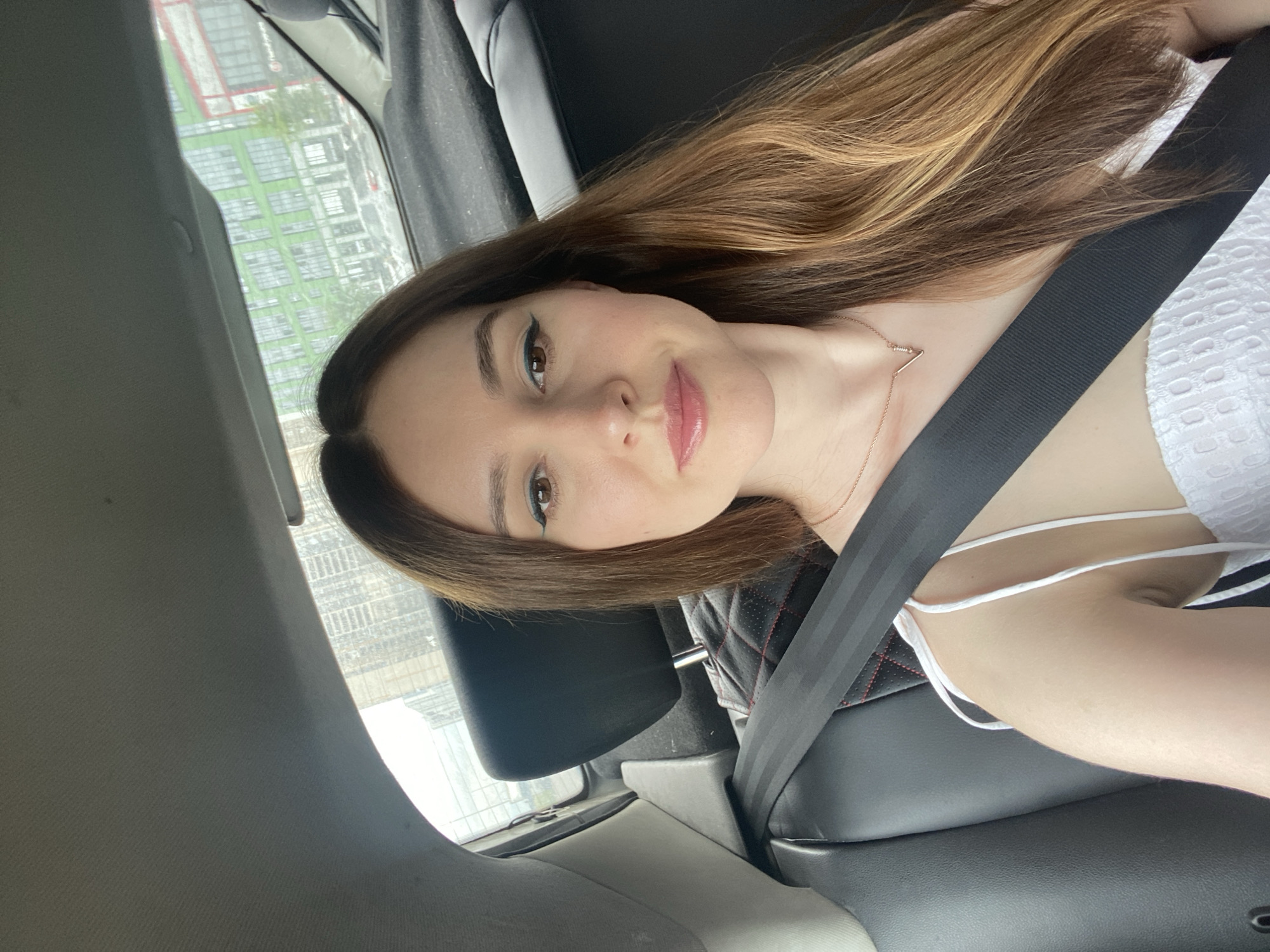 A young woman with long, brown and blonde hair is sitting in the passenger seat of a car. She is wearing a white, sleeveless dress with a textured pattern and a simple rose gold necklace. A black seatbelt is fastened across her chest. Her makeup includes winged eyeliner and glossy pink lipstick. The woman is looking at the camera with a slight smile. Through the car window, a blurred view of buildings and trees is visible, suggesting an urban or suburban setting. The lighting indicates it is daytime. The overall mood is casual and relaxed. The location is identified as Колос Снт, Russia.