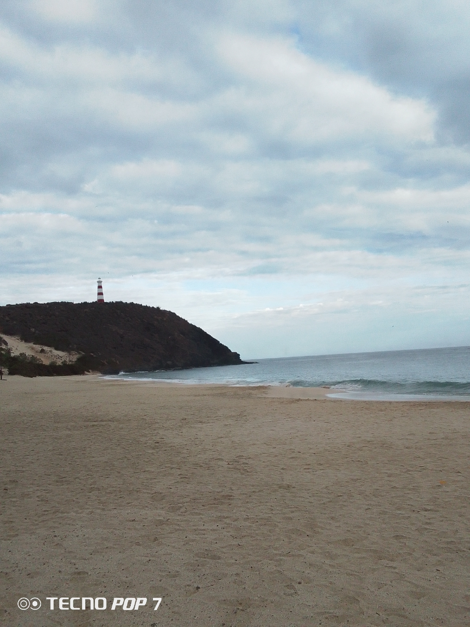 The image shows a wide sandy beach in Pedro González, Venezuela. A hill rises at the edge of the beach, topped with a red and white striped lighthouse. The sea is visible to the right of the hill, with gentle waves approaching the shore. The sky is overcast with grey and white clouds. A small amount of vegetation is visible at the base of the hill.