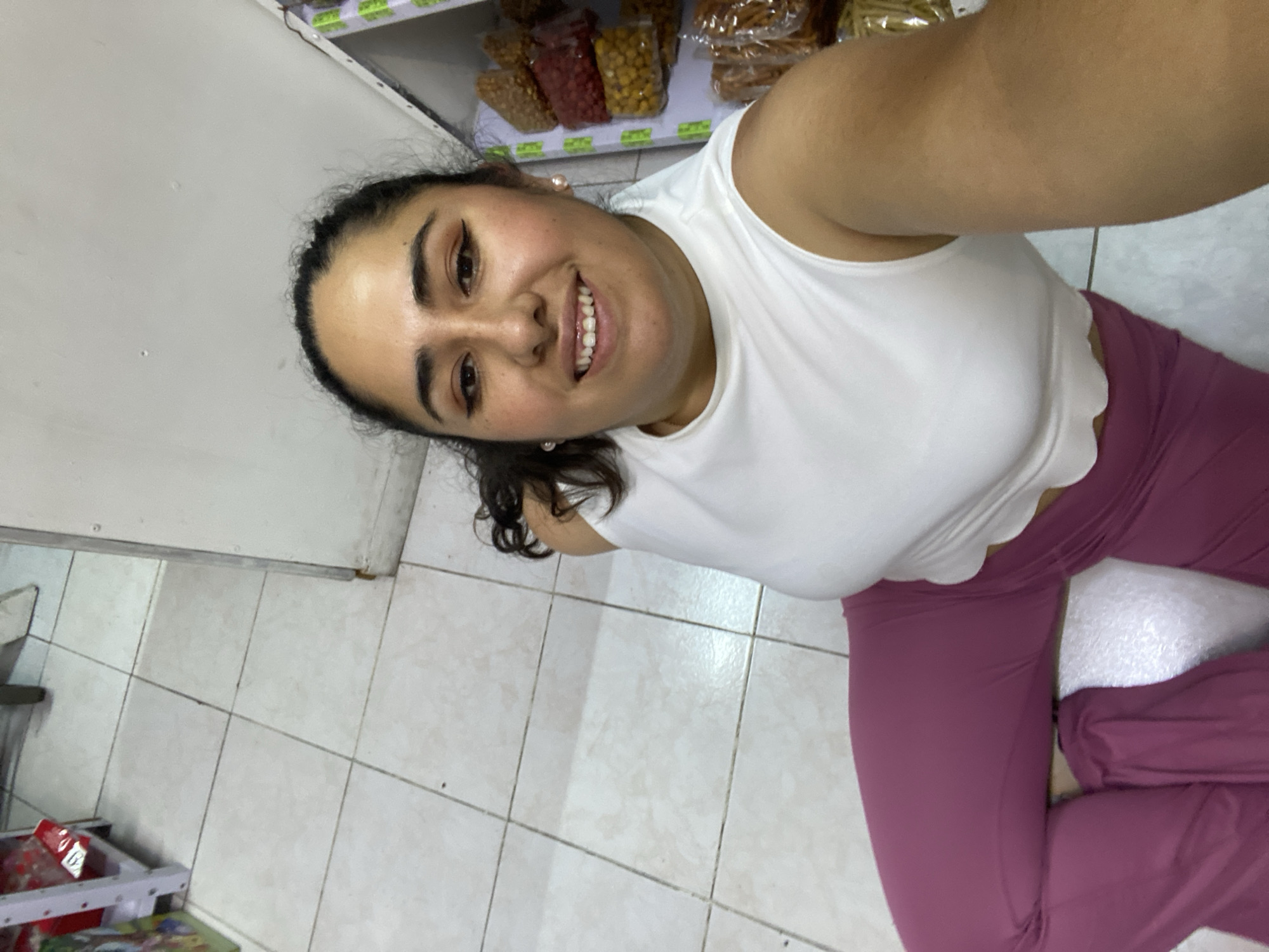 A smiling woman with dark hair and fair skin is taking a selfie in what appears to be a small shop in San Bernardino Tlaxcalancingo, Mexico. She is wearing a white tank top and pink leggings. Behind her, a shelf is stocked with packaged food items. The floor is tiled in white.