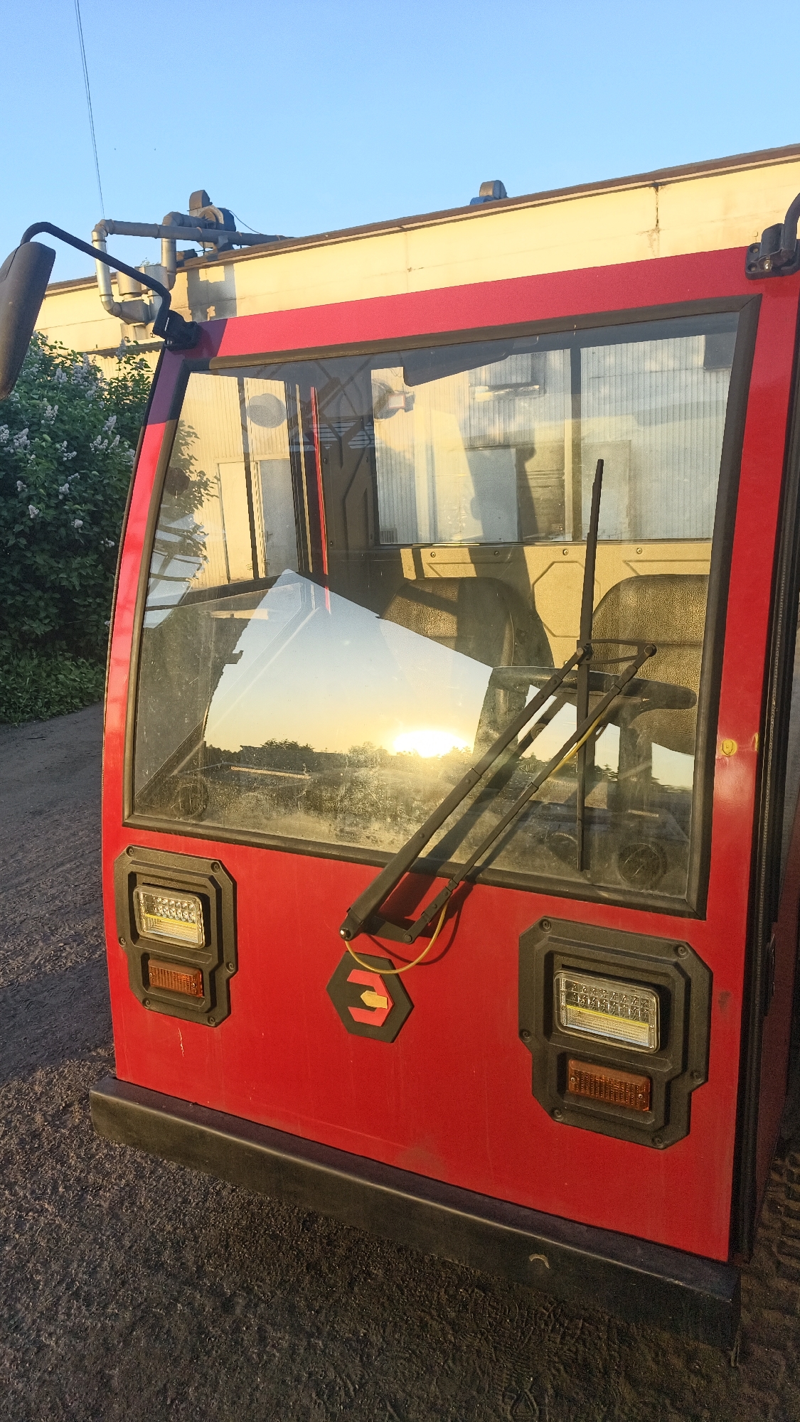 A low-angle shot shows the red cabin of a vehicle, possibly agricultural or industrial, parked outdoors in Noviy, Russia. The windshield reflects the setting or rising sun, obscuring the vehicle's interior but revealing a rural landscape on the horizon. The wipers are visible on the windshield. Below the windshield, on the vehicle's front, are two rectangular lights, each with a smaller amber light beneath. Between the lights is a hexagonal logo featuring a stylized arrow. The vehicle has a black bumper and sits on a surface of dark gravel or dirt. To the left of the vehicle, out of focus, are green bushes and a portion of a building. The sky is clear and light blue.