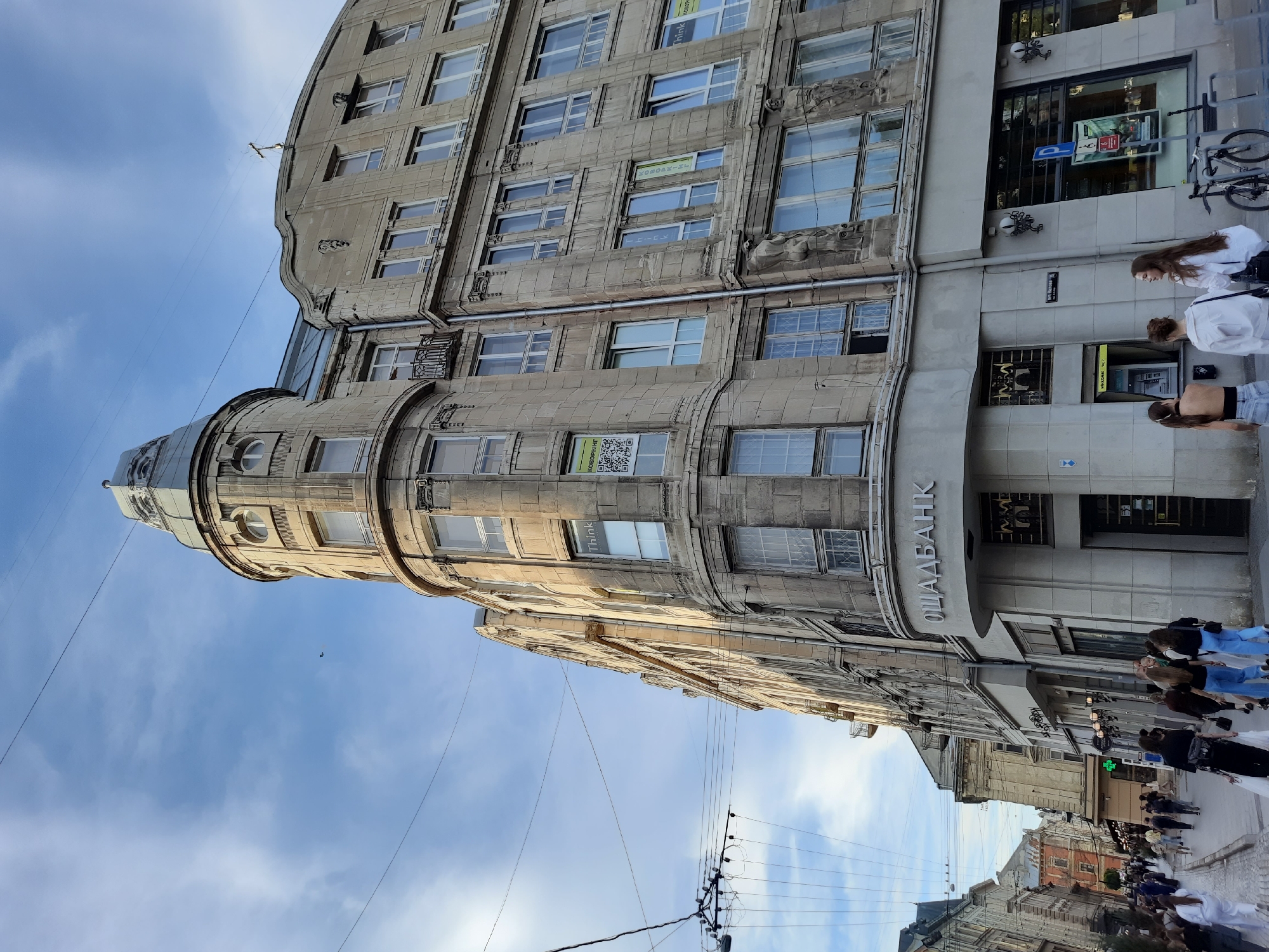 The image depicts a street view in Lviv, Ukraine, focusing on a prominent building with a curved facade. The building is constructed of light-colored stone and features multiple stories with numerous windows. On the corner of the building, a sign reads "ОЩАДБАНК," identifying it as a branch of Oschadbank. Several people are walking on the sidewalk and street in front of the building. Some are walking towards the viewer, while others are walking away. There are also utility poles and wires visible overhead. The sky is partly cloudy. The architecture suggests an older, possibly European, urban environment.