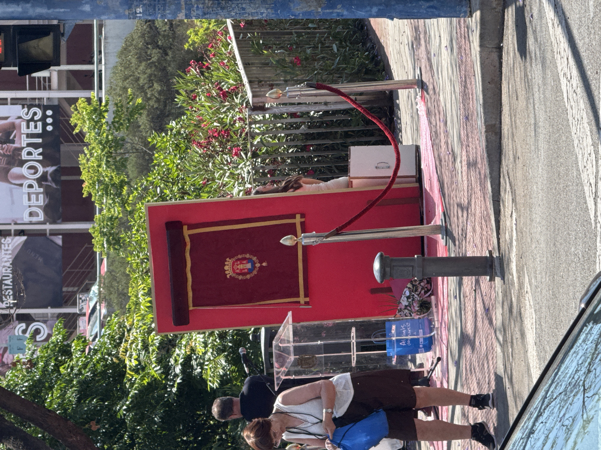 The image captures an outdoor public event or display in an urban setting during bright daylight. The scene is dominated by a red platform or stage, framed by a red velvet rope with gold stanchions, indicating a formal or restricted area. On this platform, a figure, resembling a mannequin or statue, lies partially covered by a white cloth, with long dark hair visible. Behind it, a vibrant green garden with red flowering bushes provides a natural backdrop. A burgundy banner with a golden and blue crest or coat of arms is prominently displayed on the red platform.
In front of the platform stands a clear acrylic podium with a microphone. Near the podium, a man in a dark shirt is visible, suggesting involvement in the event, possibly setting up or managing sound equipment. In the foreground, a woman wearing a white top and dark skirt, carrying a blue bag, observes the scene, facing away from the camera. Another person is partially visible beside her.
The background features urban elements: buildings with various signs, including a large sign that clearly reads "DEPORTES" (Spanish for "Sports") and another partially visible as "RESTAURANTES" (Spanish for "Restaurants"). A traffic light showing a red signal is also visible in the upper left. The ground consists of concrete pavement, partly adorned with a red patterned surface, beside an asphalt road. A portion of a car is visible on the far right. The blue bag near the podium has some text that appears to be "Alcor c co mercio". The overall setup suggests a public ceremony, unveiling, or art installation, possibly with a somber or significant theme.