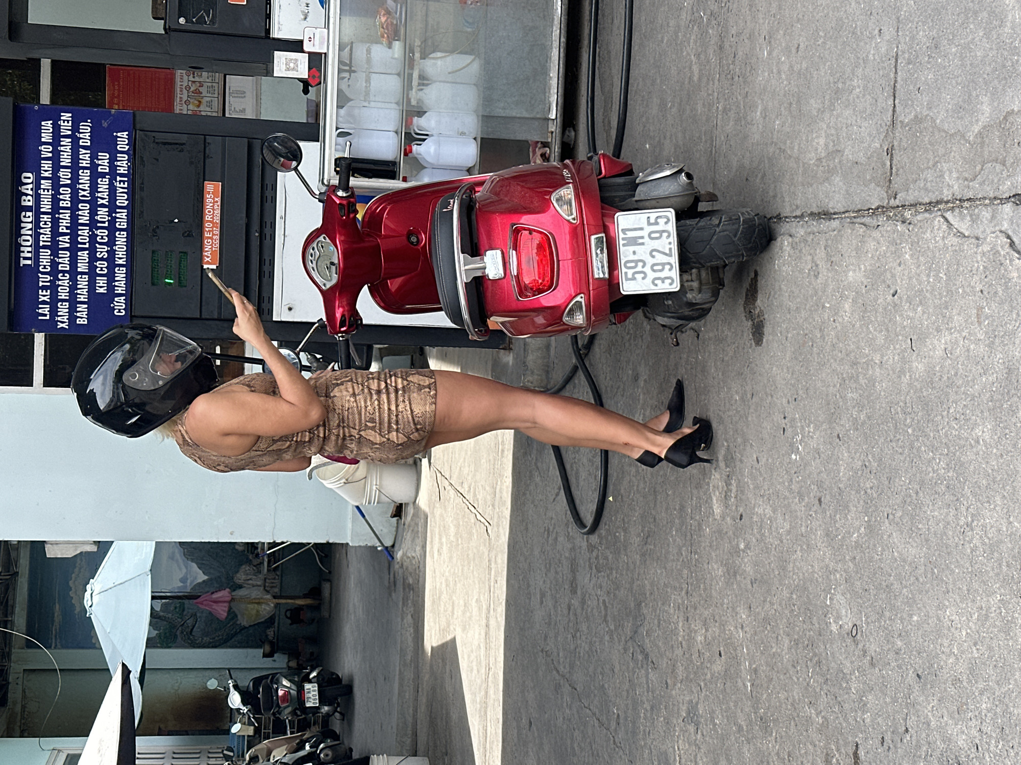 In Bac Nha Trang Ward, Vietnam, a woman wearing a snake-print dress and black high heels stands beside a red Vespa scooter at a gas station. She has a black helmet on and is looking at her phone. The scooter's license plate reads "59-M1 392.95." Behind the scooter, a gas pump displays information about E10 RON95-III fuel. In the background, other scooters are parked under an awning, and a mural of a mountainous landscape is visible on a wall. The ground is paved with concrete, and a black hose lies on the ground.