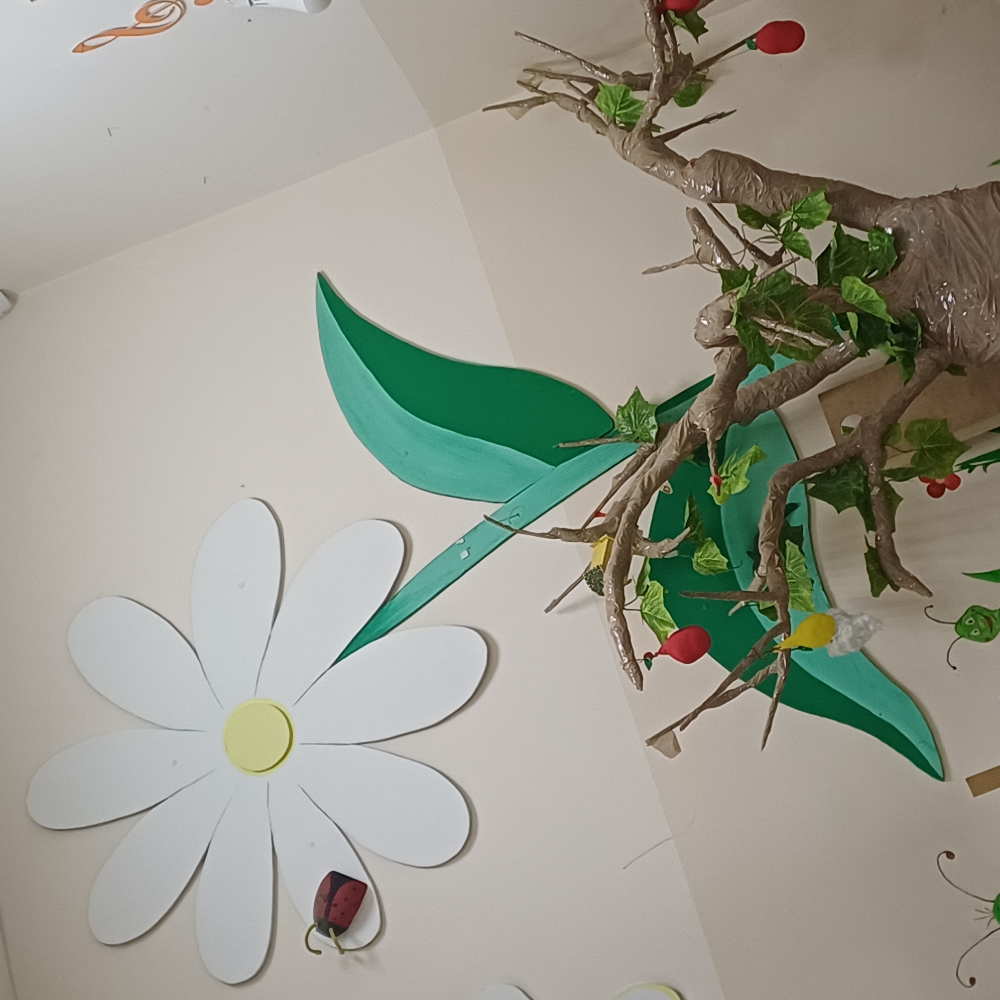 An indoor scene depicts a wall decorated with a large white daisy with a yellow center and a ladybug. To the right, a large green leaf-shaped cutout is attached to a tree trunk made of branches wrapped in brown paper. The tree has green leaves and is adorned with red and yellow fruit-shaped ornaments and a white fluffy object. Several other decorative elements, including small bugs and a musical note, are visible on the wall. The lighting is diffuse, suggesting an artificial light source.