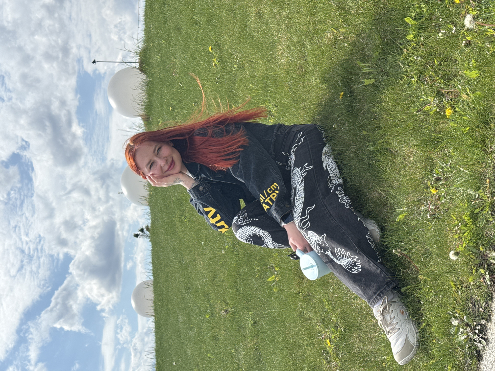 A young woman with red hair is reclining on a grassy slope under a partly cloudy sky. She is resting her head on her hand and smiling slightly towards the camera. She is wearing a black denim jacket and matching pants, both with white dragon designs, along with white sneakers. She holds a blue water bottle with a straw. In the background are white spherical objects, possibly decorative installations or art pieces. While the image does not provide enough information to confirm the precise location in Rostov-on-Don, Russia, the setting suggests a public park or landscaped area within the city.
