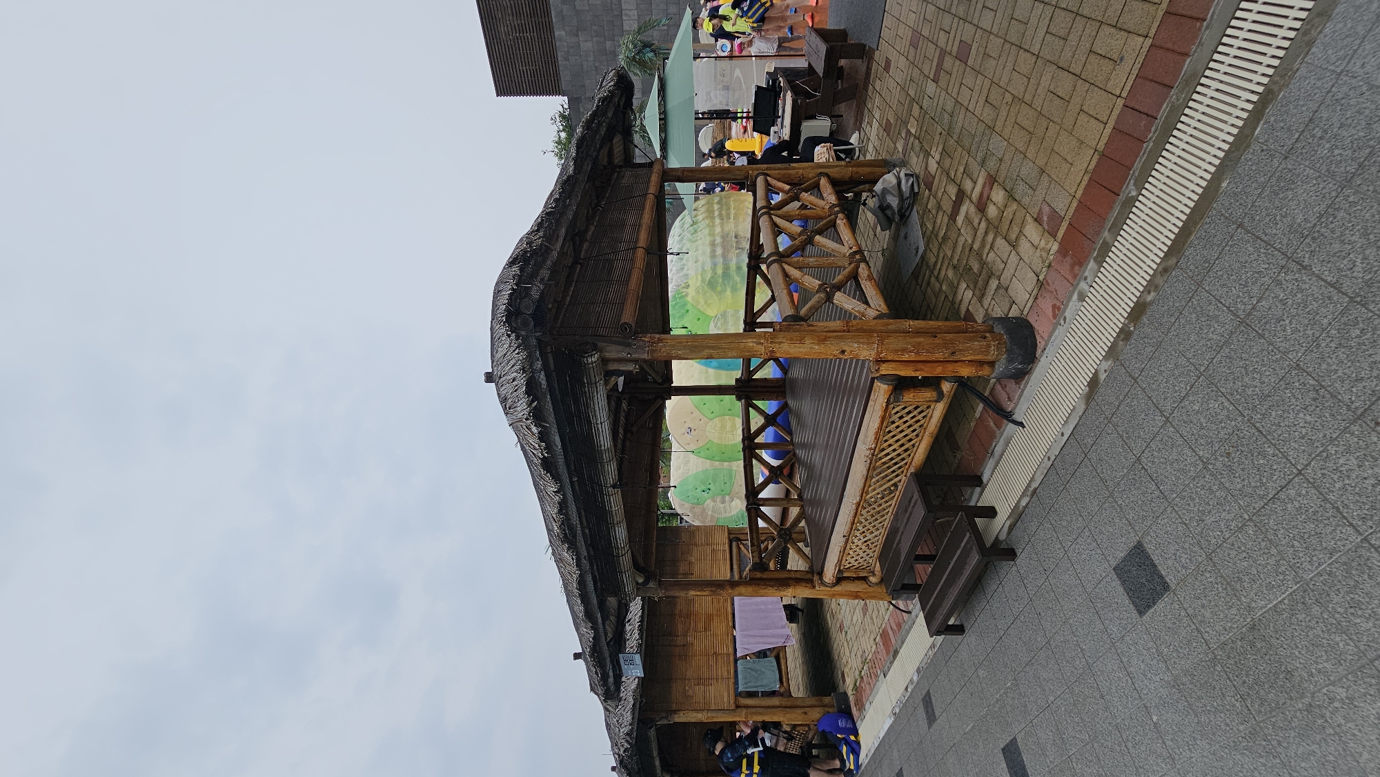 The image captures an outdoor scene at an aquatic facility in Asan-si, South Korea. In the foreground, a paved walkway with gray tiles leads to a thatched-roof wooden pavilion. The pavilion features a raised wooden deck and a decorative lattice railing made of bamboo. Two dark wooden benches are positioned on steps leading up to the pavilion. To the left of the main pavilion, a similar, smaller structure is visible, with towels hanging inside and a person wearing a life vest and shorts standing to the left. In the background, behind the pavilions, a large inflatable water toy with green and white circular patterns is partially visible. Further to the right, a green umbrella shades a table with people gathered around it, suggesting a seating or service area. The overall environment appears to be a recreational area, possibly a water park or resort, with a cloudy sky overhead.