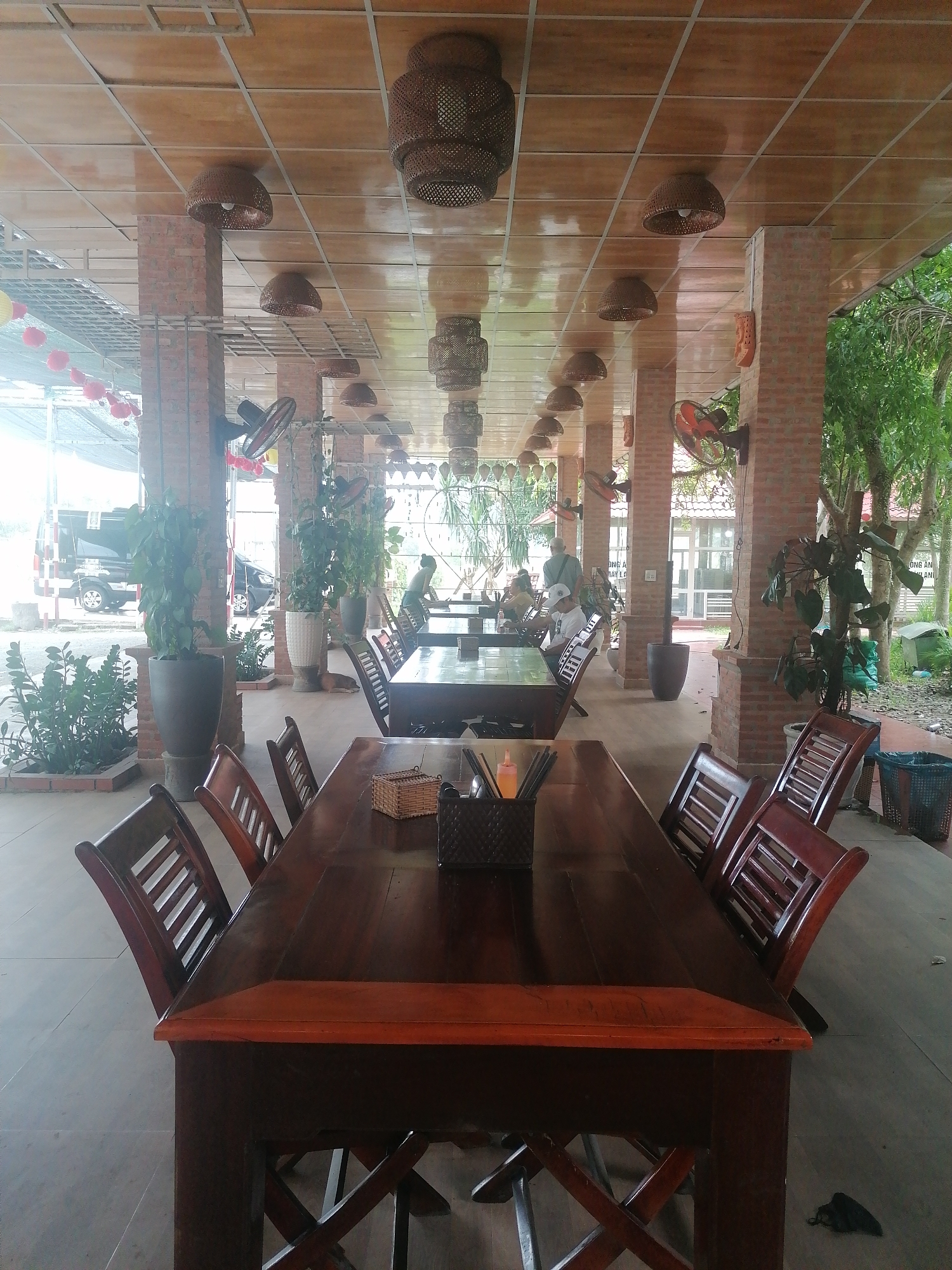 The image shows a restaurant in Nam Khanh Vinh, Vietnam. The interior is spacious, with a high ceiling made of wooden panels. Wicker lamps hang from the ceiling at regular intervals. Brick pillars support the roof. Seated in the restaurant are tables with chairs, and some plants. The building is partly open-air, with views of a garden and a street visible in the background. The scene appears to be during daylight.