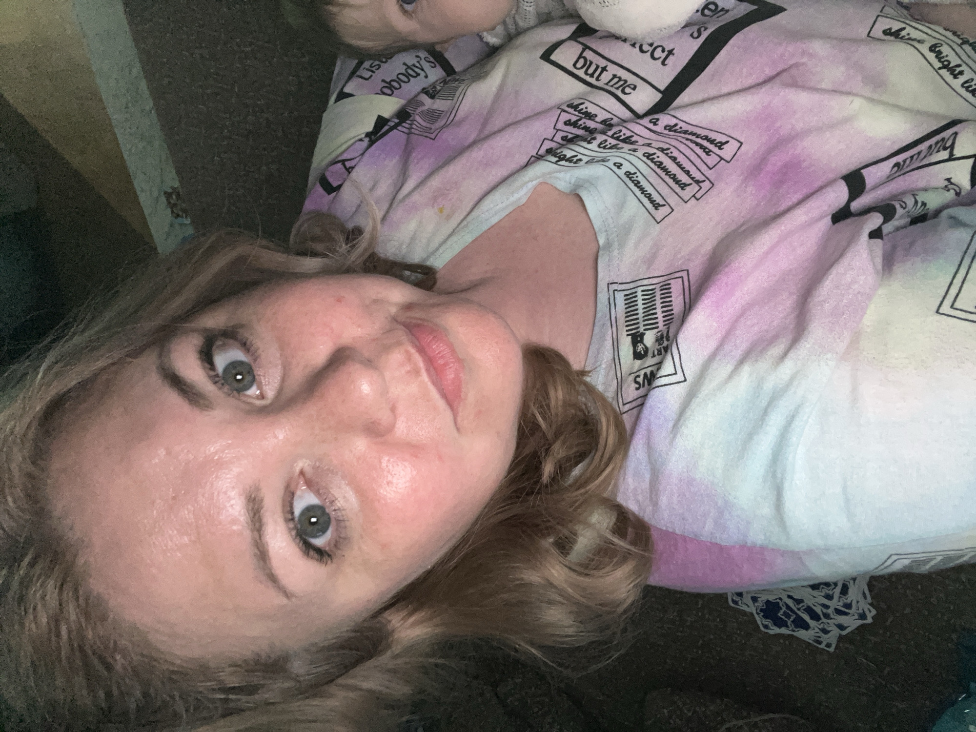 A woman is lying down, her face close to the camera. She has fair skin, light brown curly hair, and grey eyes. A baby, only partially visible, lies above her head. The woman is wearing a light-colored t-shirt with a tie-dye pattern and black lettering. The background is a brown textured surface. The OCR mentions "Lis nobody's but me" and "shine like a diamond" suggesting the shirt has graphic text on it. There is nothing in the image to suggest it was taken in Vesyoloye, Russia.