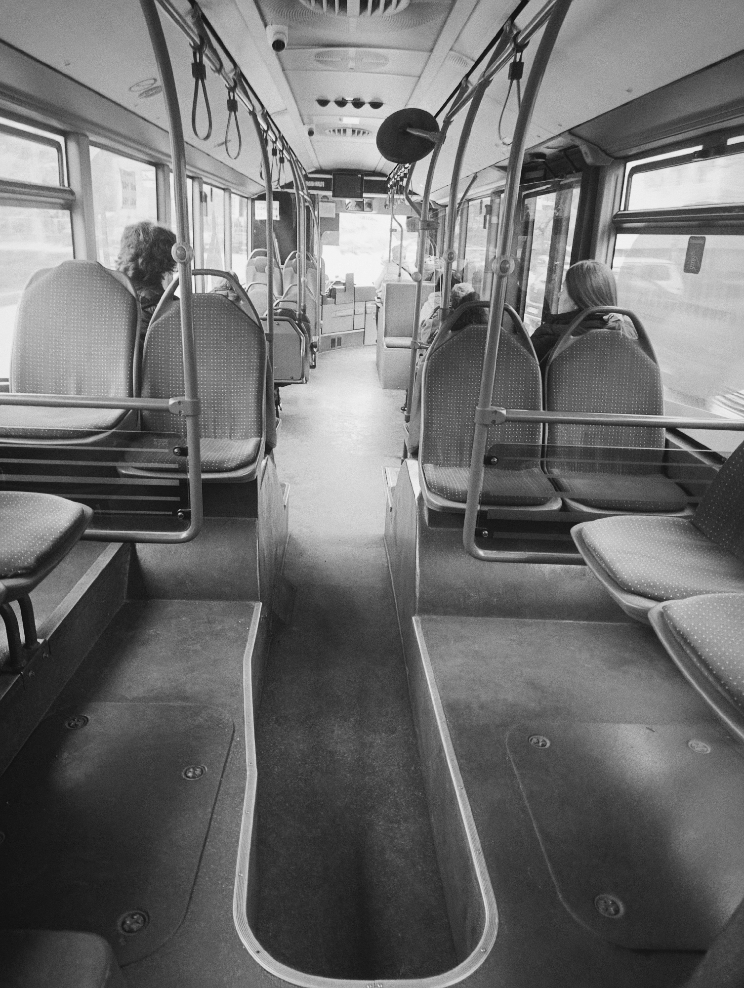 The image depicts the interior of a bus. The scene is rendered in black and white. Rows of seats with patterned upholstery line the aisle, mostly unoccupied. The floor of the bus has recessed areas alongside the seats. Hand straps hang from the ceiling. Passengers are visible in the background, including one with shoulder-length hair and another with curly hair. A security camera is mounted on the ceiling. The background windows reveal exterior views, but the location is not discernable as Kardzhali, Bulgaria.
