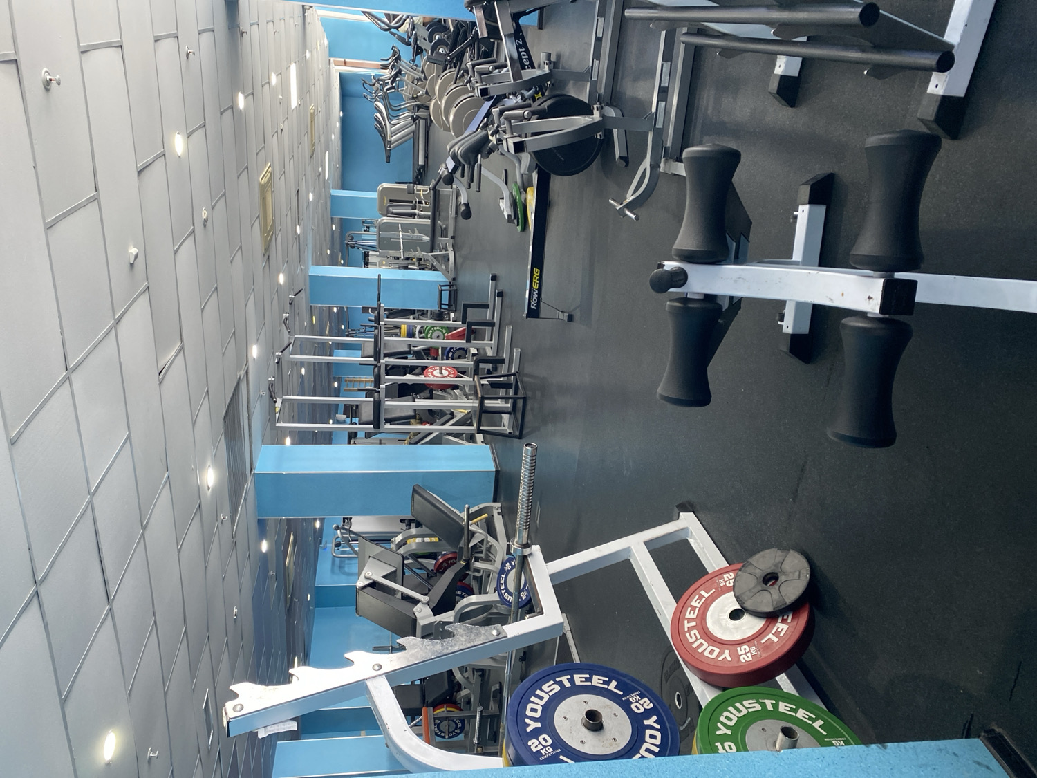 This image depicts the interior of a well-equipped gym or fitness center, rotated 90 degrees counter-clockwise. No people are present, and the equipment appears ready for use.
In the foreground and midground, various strength training machines are visible. A leg press machine is prominent, surrounded by several "YOUSTEEL" brand weight plates. These plates come in different colors and weights, including blue (20 KG), red (25 KG), and green (10, likely 10 KG). Another piece of equipment, possibly a squat rack or a multi-functional strength station with black padded rollers, is also clearly seen.
Further into the gym, a "ROWERG concept 2" rowing machine is identifiable. Beyond this, numerous other exercise machines, including additional weight stations and cardio equipment, line the space, though they are less distinctly visible in the background. The floor is dark, suggesting rubber matting common in gyms. The ceiling consists of light gray square tiles with recessed circular lights. Light blue structural elements, possibly pillars or wall sections, are integrated throughout the gym, adding a contrasting color to the mostly metallic equipment and dark floor. The overall setting suggests a modern and functional fitness environment.