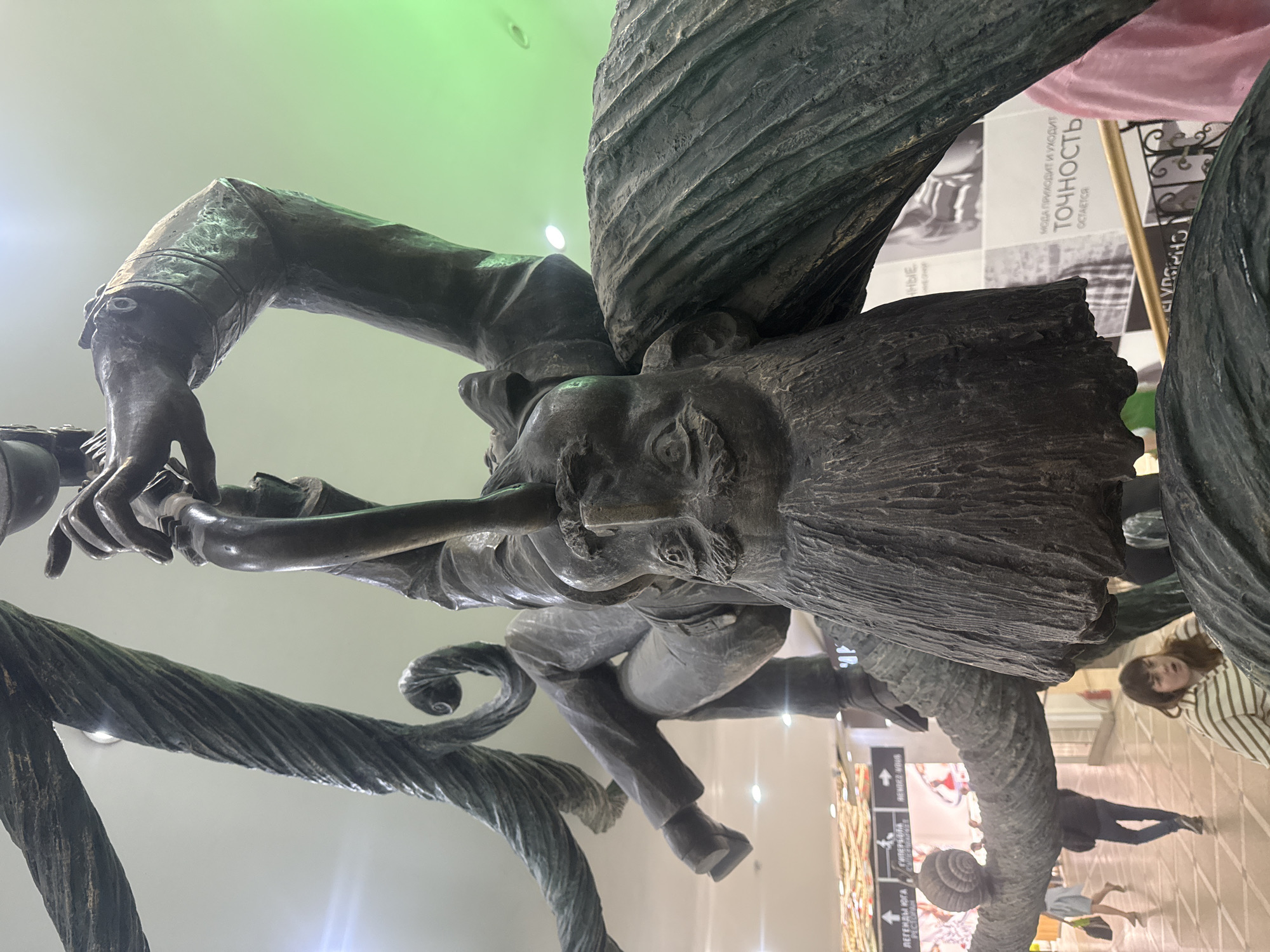 The image depicts a bronze statue of a man with long hair and a mustache, seated and playing a musical instrument that resembles a pipe or clarinet. The statue is mounted on twisting, rope-like structures. The statue is located in the city of Yekaterinburg, Russia. In the background, there are commercial displays with text in Russian and people reflected in the shiny floor. There are also architectural elements of the building, including walls and possibly parts of the ceiling. A young woman with light brown hair, wearing a horizontally striped top, is also visible in the background.