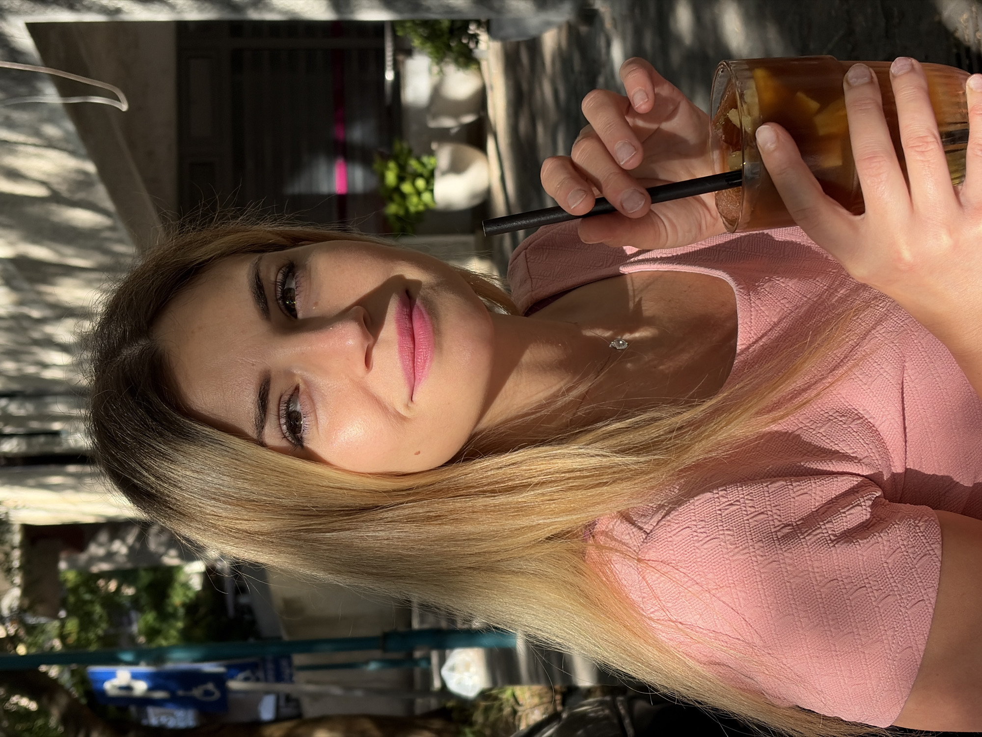 A young woman with blonde hair and pink lipstick holds a glass of iced coffee in her right hand, resting her left hand on top of the glass. She is wearing a pink textured top and a delicate necklace. The scene appears to be outdoors, possibly at a cafe or restaurant, with sunlight casting shadows on her face and the background. In the background, there are potted plants, a building with large windows, and a blue parking sign. The overall impression is a casual, sunlit moment.