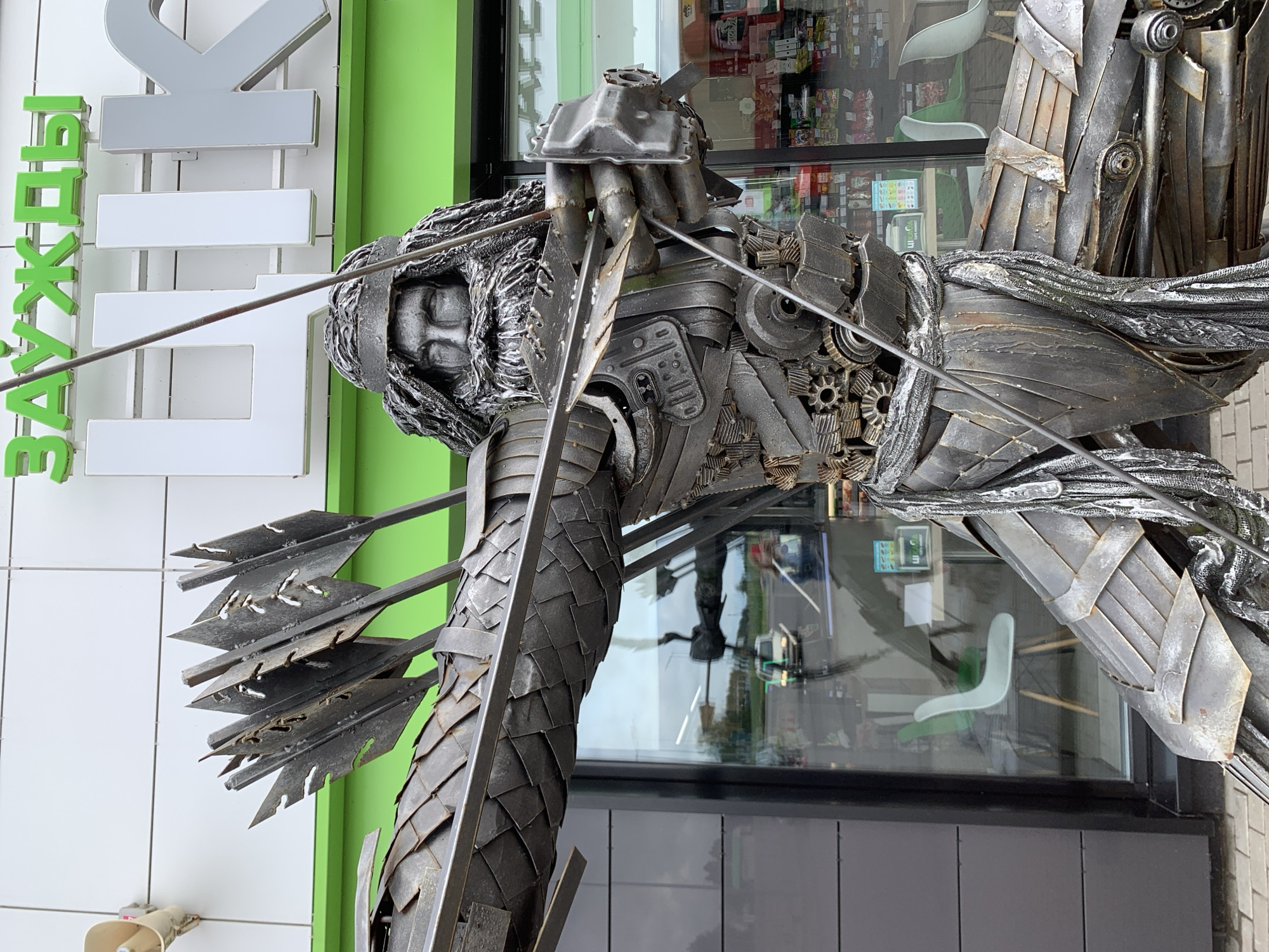 The image shows a metal sculpture of a medieval archer standing outside a store with a green awning. The sculpture is made of various metal parts, including gears, plates, and pipes, and depicts the archer in a dynamic pose, drawing a bow with arrows in his quiver. The store's facade features large windows and a sign with Cyrillic lettering. In the background, through the windows, shelves stocked with goods and chairs are visible, suggesting an interior retail space. The overall setting appears to be an urban or suburban environment, possibly a commercial area. The location is identified as Дзешчанскі сельскі Савет, Belarus, implying the sculpture is a public art installation in this region.