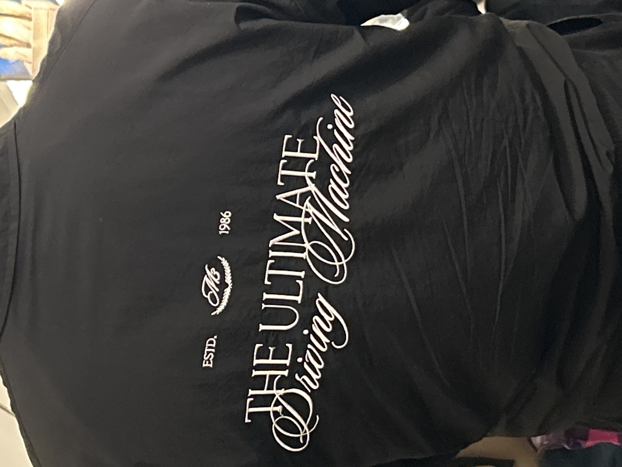 A close-up, slightly angled shot shows the back of a black garment, likely a jacket or shirt, featuring white printed text and a logo. The fabric appears somewhat wrinkled, suggesting it might be hanging or draped.
The prominent text reads "THE ULTIMATE Driving Machine." "THE ULTIMATE" is in a sans-serif, uppercase font, while "Driving Machine" is in an elegant, flowing script font, with the "D" and "M" being particularly ornate.
Above and to the left of "THE ULTIMATE," there's a stylized logo that appears to be the letters "Mb" or "M&b" enclosed within a laurel wreath design. To the left of this logo, the word "ESTD." is printed in a simple, uppercase font. To the right of the logo, the year "1986" is printed in the same simple, uppercase font.
The background is out of focus, showing indistinct shapes and colors, consistent with an indoor setting. The lighting is even, suggesting artificial indoor light. No individuals are visible in the frame. The image focuses entirely on the details of the black garment and its white embellishments.