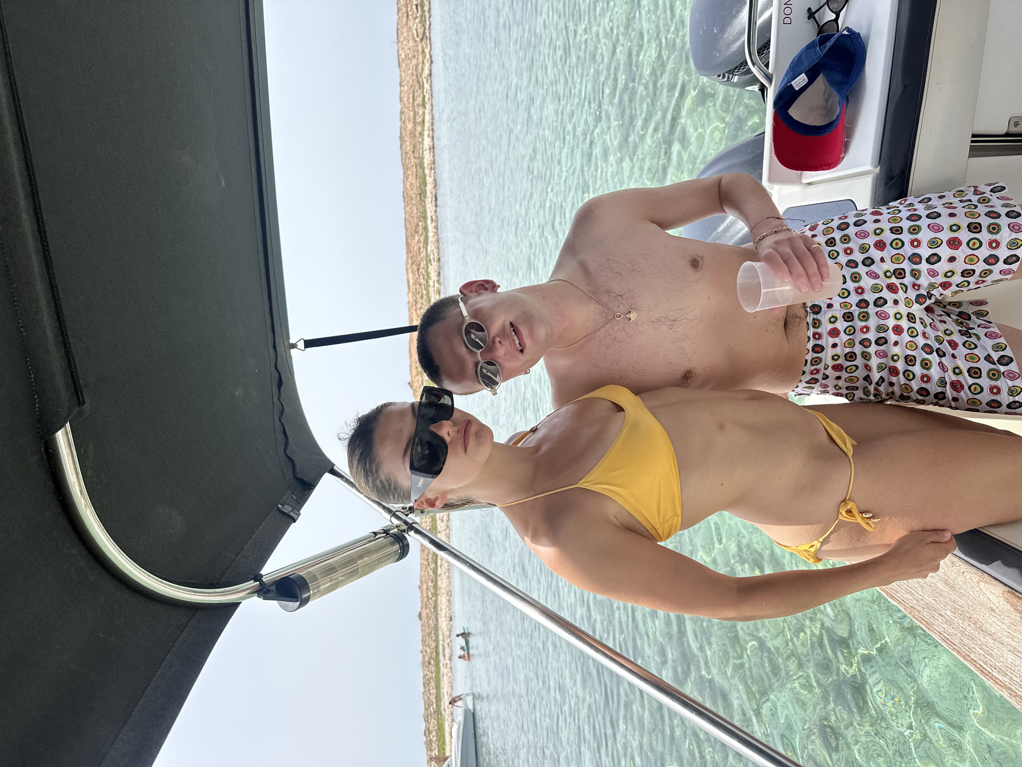 A medium shot shows a man and a woman standing on a boat with clear turquoise water and a rocky coastline in the background. The woman, on the left, wears a yellow bikini and large black sunglasses. The man, on the right, wears patterned swim trunks and sunglasses. He holds a plastic cup in his right hand. A red and blue baseball cap and a pair of sunglasses rest on the boat's console. The sky is overcast.
