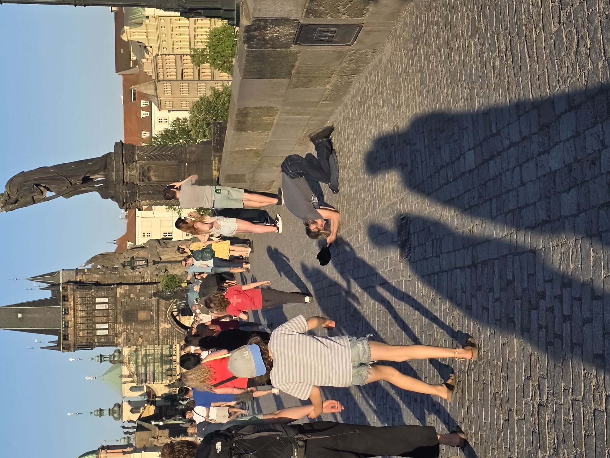 This image depicts a crowded scene on Charles Bridge in Prague. Pedestrians, mostly tourists, are spread across the cobblestone walkway. Some are standing near a statue on the bridge, while others are walking or standing in groups. A man appears to be in the middle of a handstand. The shadows of the people are visible on the cobblestones. The Old Town Bridge Tower and various buildings in the city are visible in the background under a clear blue sky.