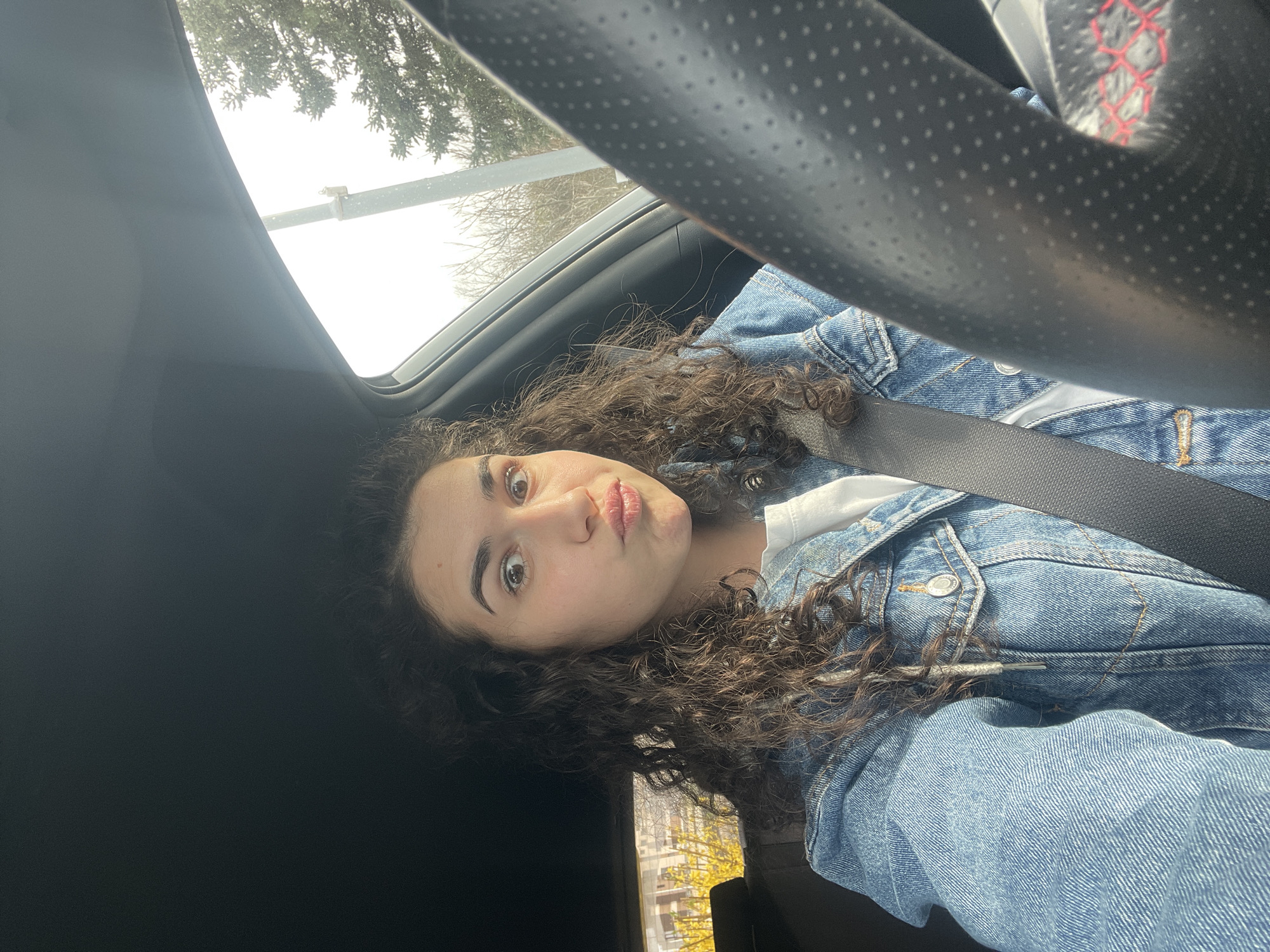 A young woman is captured in a playful selfie taken inside a car, likely in Sofia, Bulgaria. She is positioned on the right side of the frame, looking directly at the camera with wide, light brown eyes and a puckered-lip expression. Her dark, curly hair is prominent, framing her face and cascading over her shoulders. She is wearing a light blue denim jacket over a white top, secured by a black seatbelt that crosses her chest.
The interior of the car is visible, including a dark, perforated steering wheel with hints of red stitching in the upper right. Above the woman, through the car's roofline and window, an overcast sky and the bare branches of trees are seen, suggesting cooler weather. Another window on the lower left reveals faint hints of yellow foliage, indicating an autumn season. The bright light outside confirms it is daytime. The scene conveys a casual, personal moment. There is no visible text in the image.