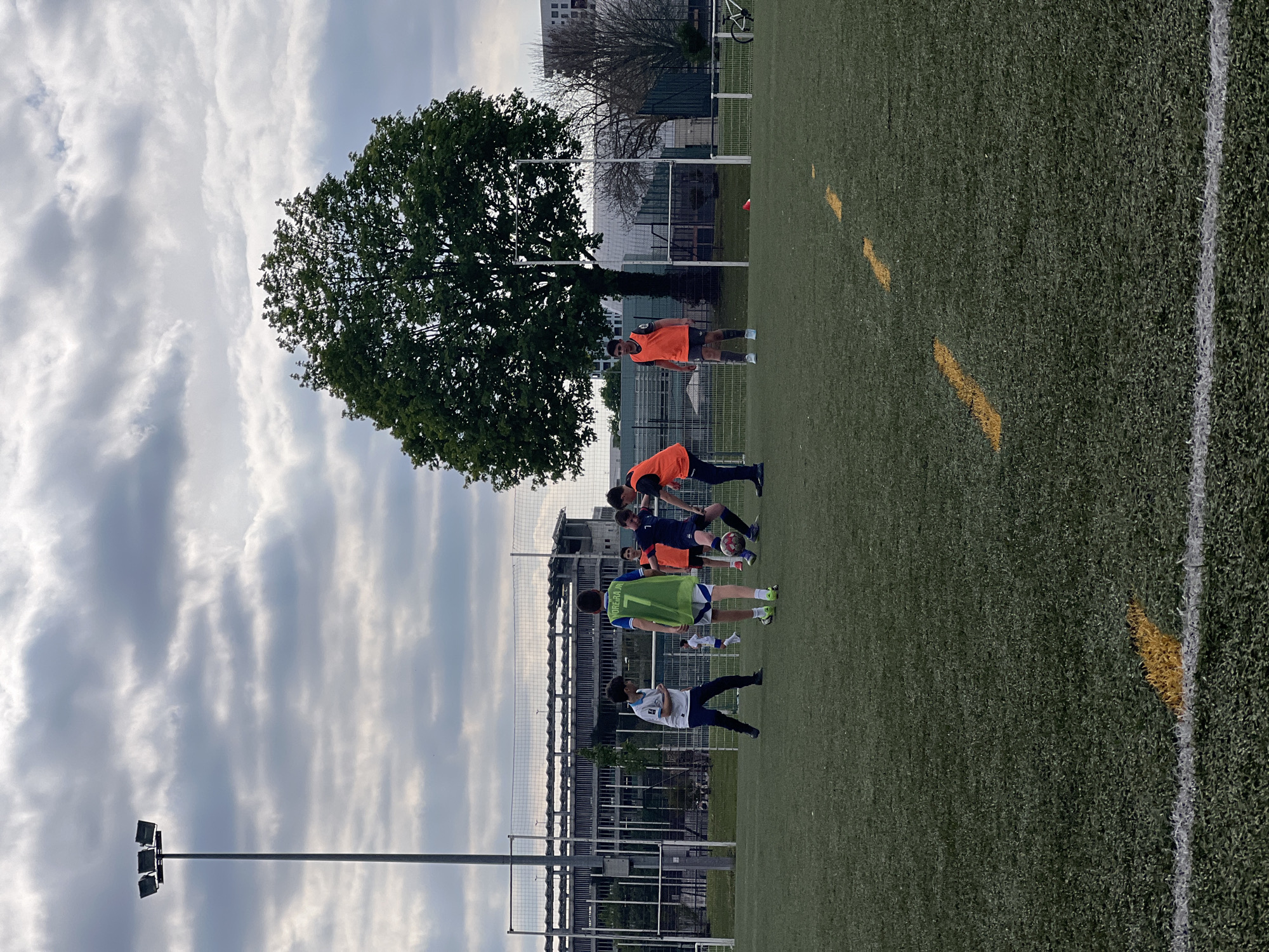 A horizontal outdoor shot shows a group of soccer players on an artificial turf field, likely in Strasbourg, France, given the architectural style. The sky is cloudy. A tall tree with dense foliage stands on the left side of the field. A row of soccer players in assorted jerseys is centered on the field, some appearing to stretch or bend over. Behind them is a chain-link fence and a building under construction. A tall light pole is situated on the far left, extending into the cloudy sky. On the right side of the field, yellow lines are visible on the green turf. In the background, a soccer goal is partially visible.