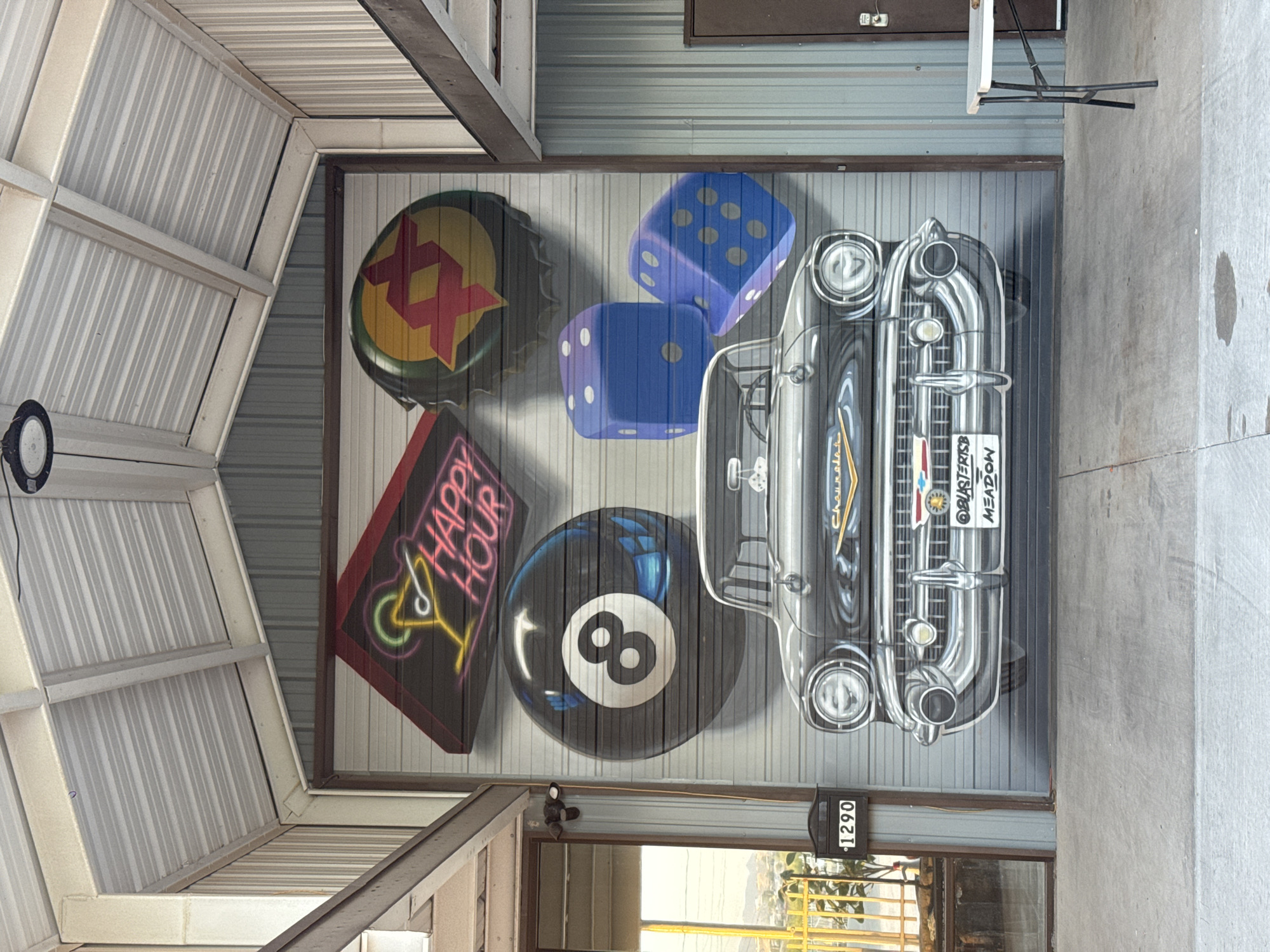 The image shows a garage door in Sunland Park, United States of America, that has been painted with a mural. The mural features a vintage black Chevrolet car in the center, with various objects arranged around it. To the top left of the car, a neon sign displays a cocktail glass and the words "HAPPY HOUR". To the top right, a large bottle cap with a red and yellow starburst design is depicted. Directly above the car, a large black eight-ball from a billiard game is painted. To the right of the eight-ball, two oversized blue dice are shown, one with four dots and the other with five dots. The garage door itself is made of corrugated metal and is painted to appear as a mural. The surrounding environment includes the roof structure of the garage, with exposed beams and corrugated metal roofing. To the left of the garage door, a glass door and a mailbox with the number "1290" are visible. A metal table with a white surface stands to the right of the garage door. The ground in front of the garage is concrete.