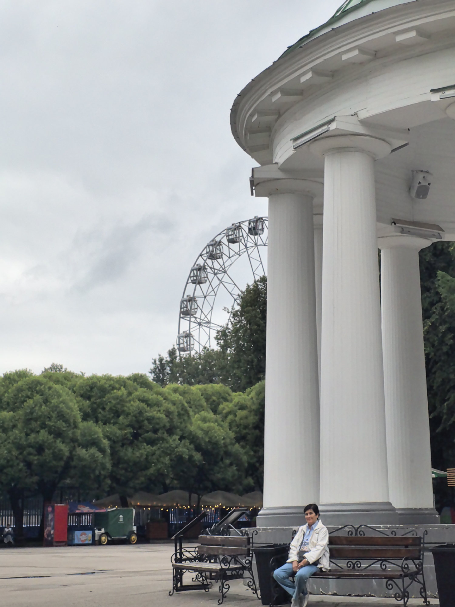In the city of Perm, Russia, a woman sits on a wooden bench in front of a large white structure with fluted columns. The woman is wearing a white jacket and blue jeans. Behind the bench are three trash cans. In the background, a Ferris wheel is visible against a cloudy sky, along with lush green trees and what appear to be food stalls or kiosks. The overall atmosphere suggests a public park or amusement area.