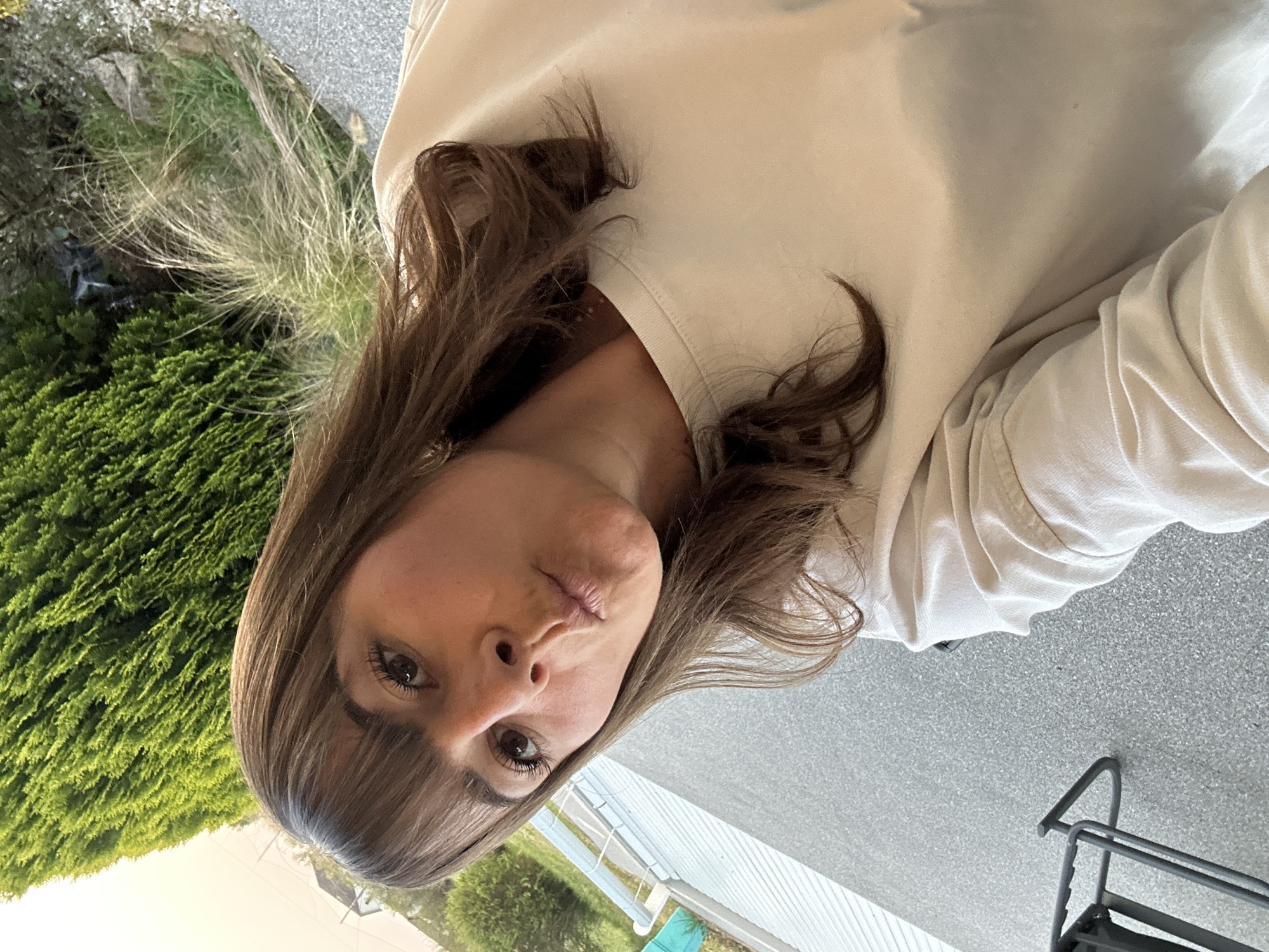 A woman with brown hair and bangs is positioned in the foreground, her face close to the camera. She has brown eyes and is making a kissing face. She is wearing a light-colored, loose-fitting shirt.
Behind her, there is a view of an outdoor scene. Dense green bushes and shrubbery are visible to the left, with patches of grass and some bare branches or twigs interspersed. A gray paved surface, possibly a driveway or sidewalk, occupies the right side of the background. Further in the distance, white fencing or siding can be seen, with glimpses of a blue object and more greenery beyond.
The location is in the city of Unknown, Unknown.