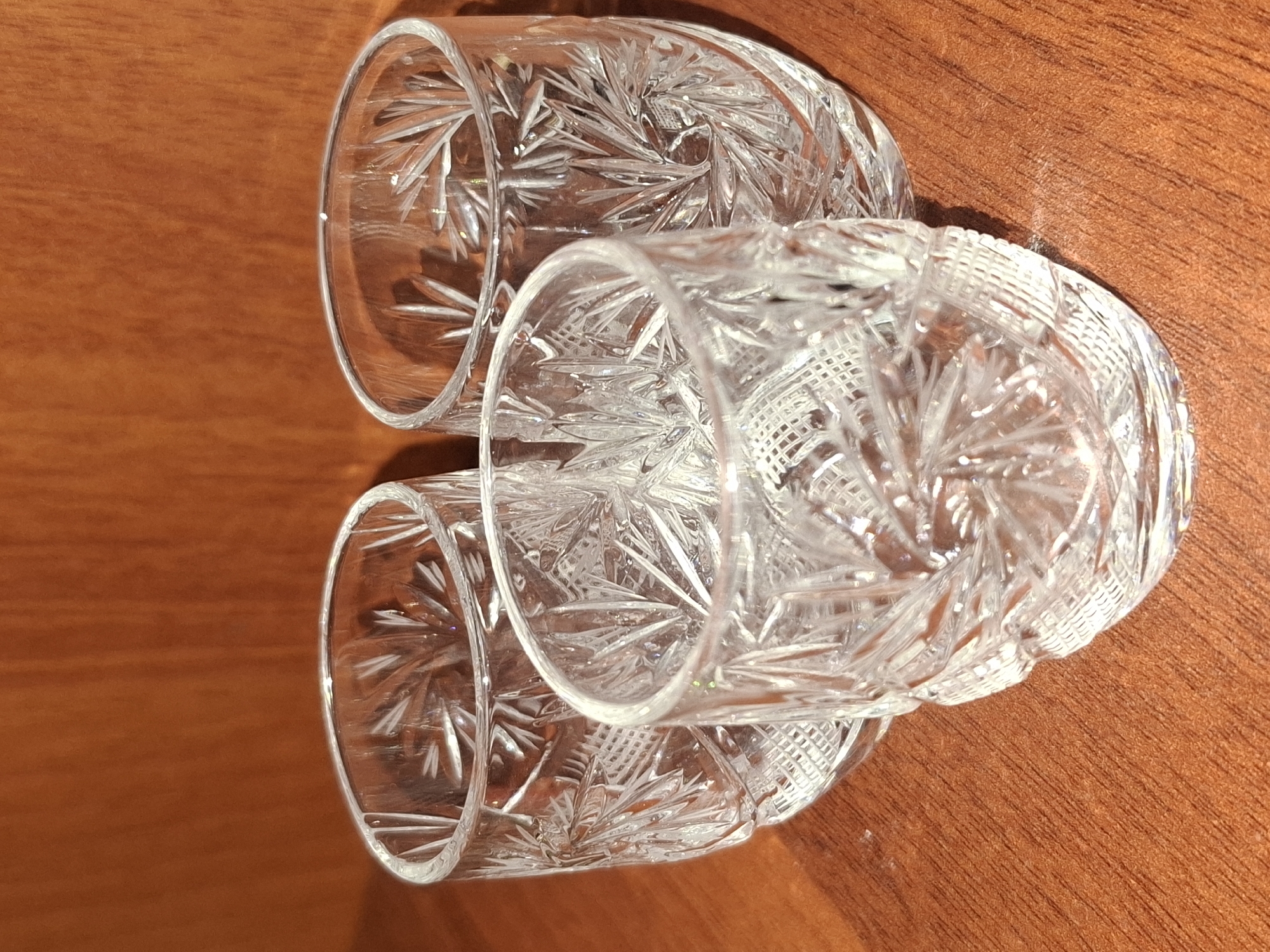 Three clear glass shot glasses with intricate starburst and diamond cut patterns are arranged on a polished wooden surface. The glasses are positioned in a triangular formation. The background consists of a textured, reddish-brown wooden surface, likely a table or shelf. The lighting casts shadows from the glasses onto the surface. There is no indication of the city of Almaty, Kazakhstan or any other specific location.