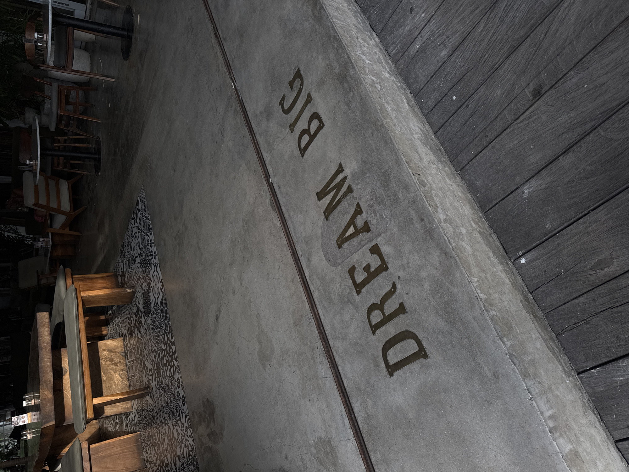 The image depicts an outdoor seating area in Kuta Selatan, Indonesia, at night. The focus is on a concrete structure with the words "DREAM BIG" etched into its surface. Adjacent to the concrete structure is a dark wooden deck. To the left, there is an area with tables and chairs, with a tile floor featuring a black and white pattern. The tables are surrounded by wooden chairs. The lighting is subdued, with shadows and variations in light intensity.