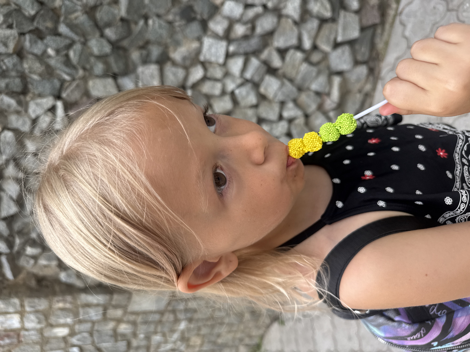 A close-up, medium shot shows a young girl with blonde hair eating a lollipop on a stick, composed of small, spherical candies in yellow and green hues. She is looking to her right, with her mouth slightly open around the lollipop. She wears a black halter top with a white patterned design and a colorful backpack straps are visible. The background is a textured wall made of grey stones. The scene appears to be outdoors, possibly in a park or street, within the city of Partenit, Ukraine.