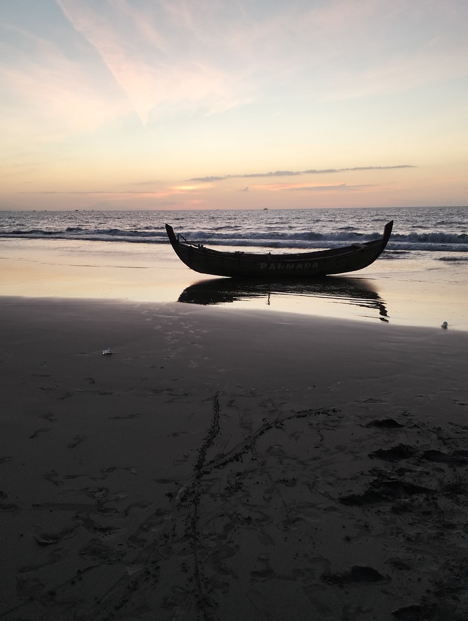 A wooden boat, with the name "PARMADA" visible on its side, rests on a wet sandy beach during twilight. The boat is angled slightly towards the right, with its bow lifted. Gentle waves lap at the shore, and the water reflects the soft hues of the sunset sky, which transitions from pale yellow and orange near the horizon to a light blue and pink higher up. Footprints are imprinted in the sand in the foreground, along with some scattered debris. The scene appears peaceful and still, characteristic of a quiet evening at the seaside. The location is specified as Uleekareung, Indonesia.