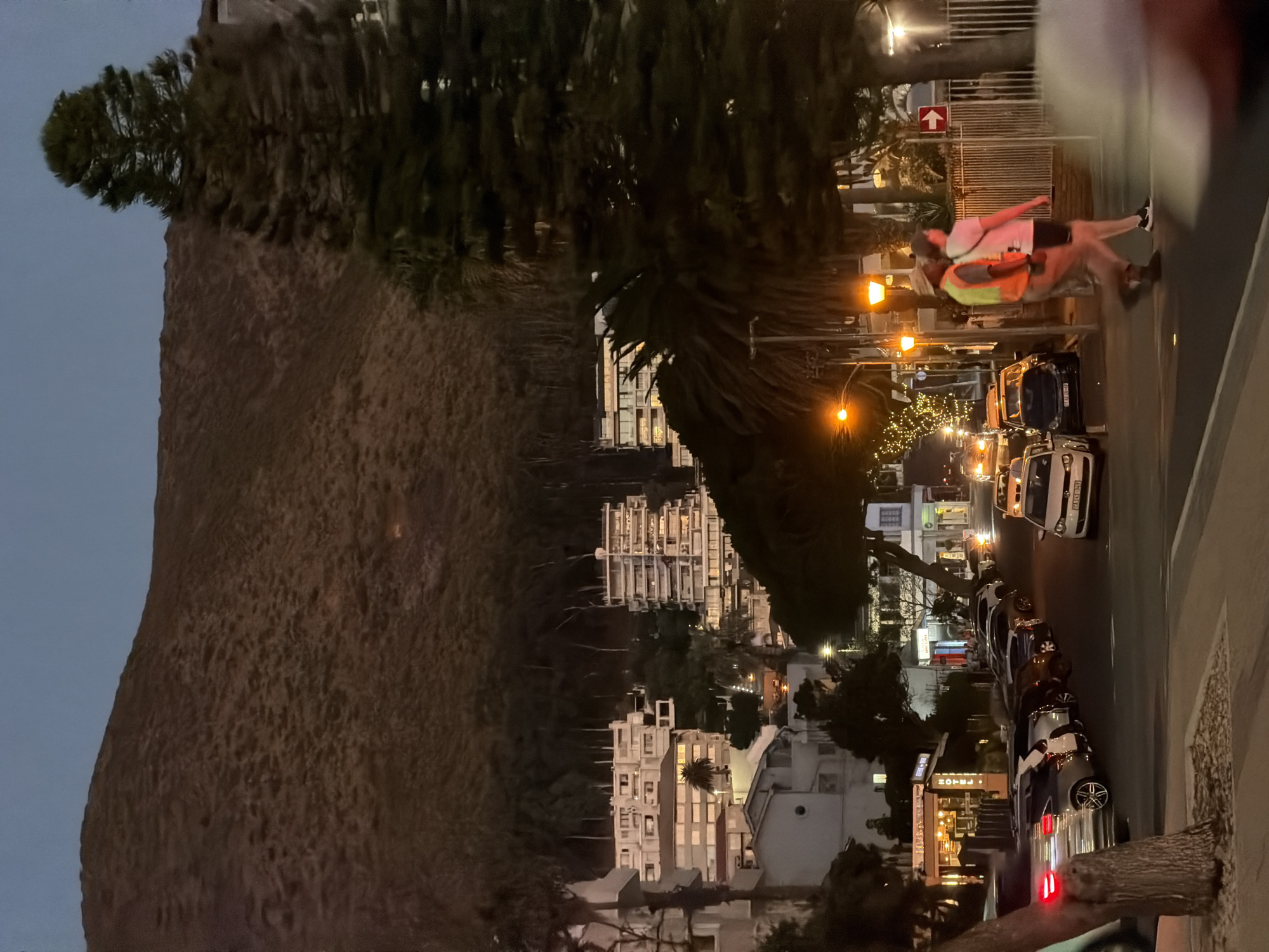 The image shows a street scene in Cape Town, South Africa at dusk. A large mountain dominates the left side of the frame. Buildings are seen in the background. A street runs along the right side of the image with parked cars. Pedestrians, one wearing a bright vest, are crossing the street. Streetlights are illuminated along the roadway.