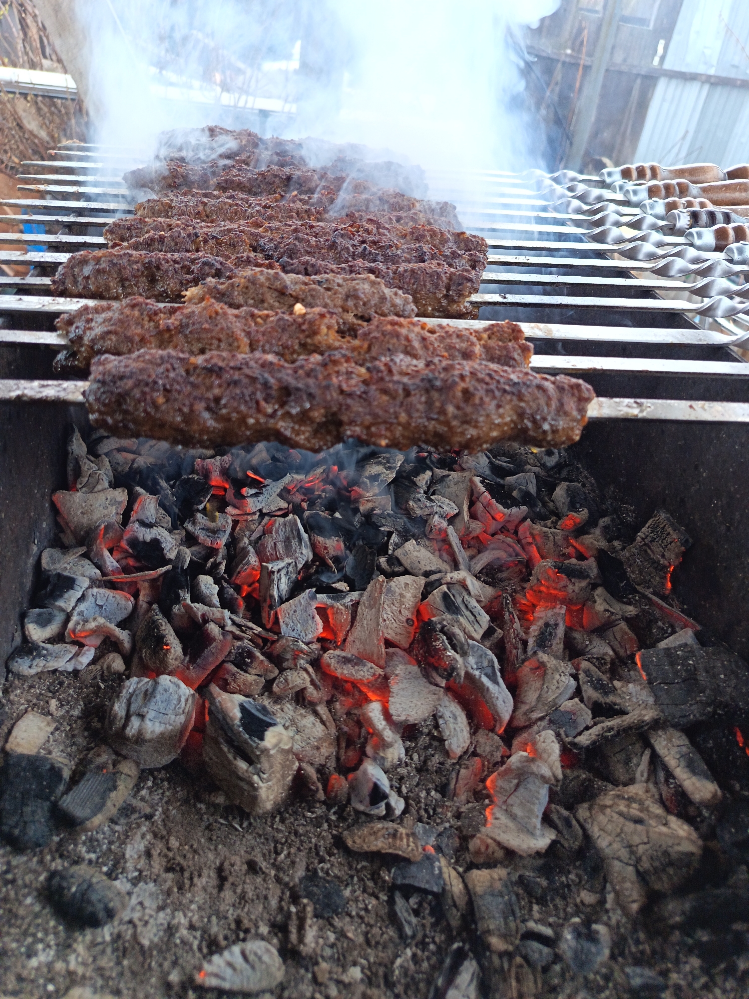 The image shows kebabs cooking on a grill over hot coals. The kebabs are on skewers, arranged in rows on the grill. Smoke rises from the kebabs and coals. The coals are glowing red in places. The photograph appears to be taken outdoors. I have no information to determine a location in Nizhnevartovsk, Russia.