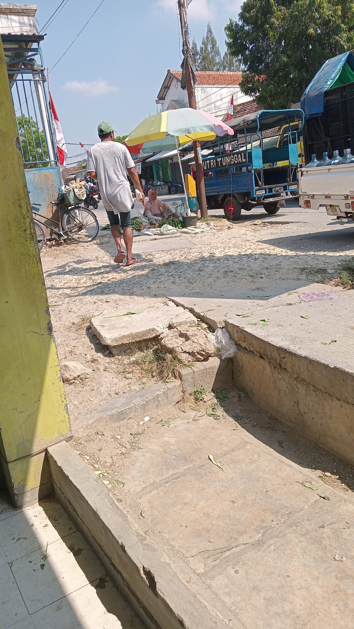 In Kunduran, Indonesia, on a bright, sunny day, a man wearing a grey t-shirt and a green cap walks away from the camera. He is on a dirt path adjacent to a paved area with a concrete channel. To his left, a bicycle is parked near a blue and white gate with an Indonesian flag. Behind him, a woman sits on the ground under a colorful umbrella, selling produce. A blue truck with the words "PUTRI TUNGGAL" is parked nearby, along with a white truck carrying several large water bottles. The environment appears to be a roadside market or informal vendor area, with buildings and trees visible in the background. The ground is a mix of dirt, gravel, and cracked concrete, indicating a somewhat uneven and weathered urban setting.