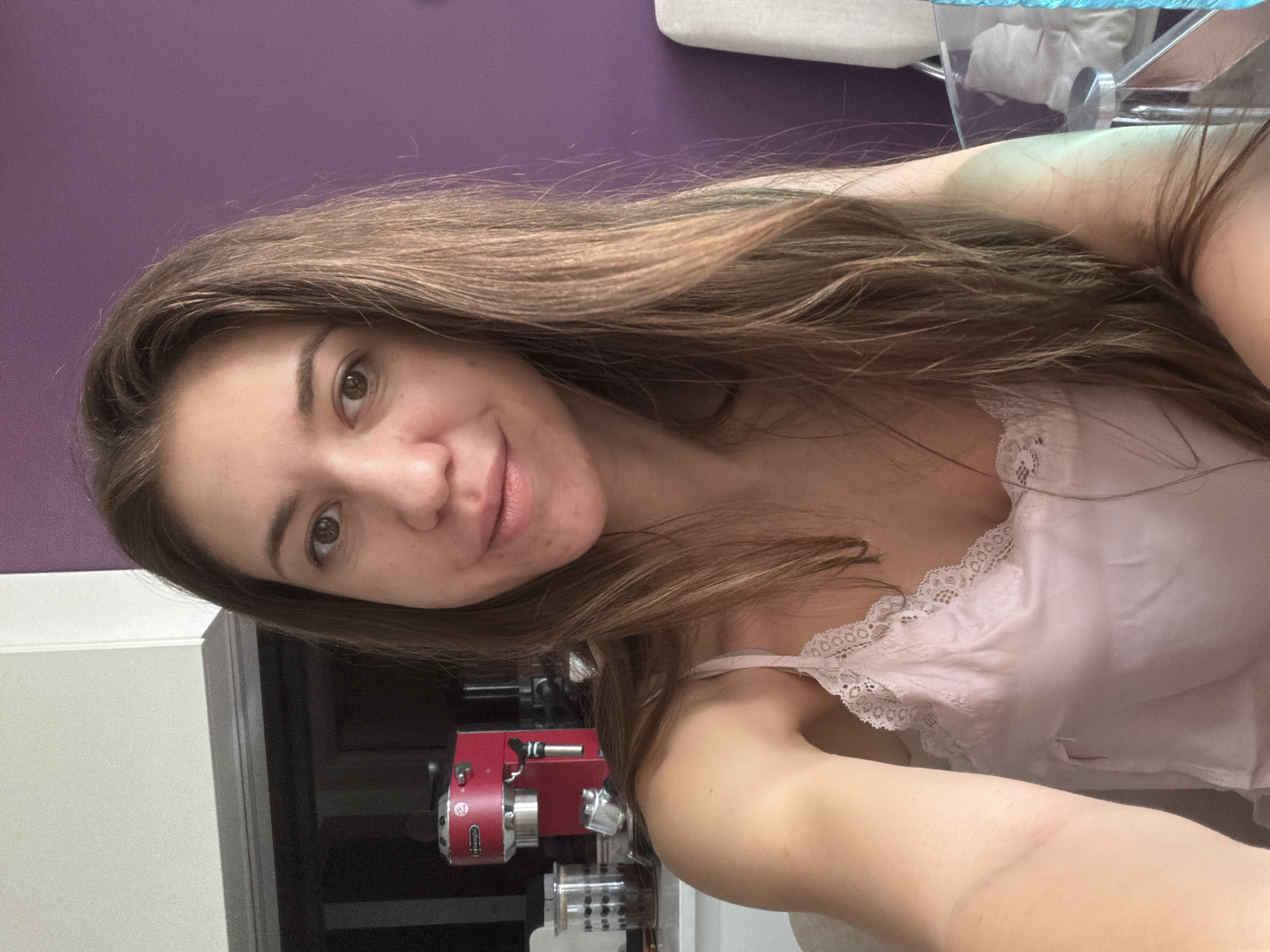 A young woman with long brown hair and light brown eyes is captured in a selfie. She is wearing a pink camisole with lace trim. Her skin appears natural with some minor blemishes. Behind her, a red espresso machine is visible on a kitchen counter, alongside a glass jar filled with coffee beans. The background features a purple wall and part of a chair. The overall setting suggests a casual indoor environment, likely a kitchen or dining area. The image does not contain any information that would place it in Sofia, Bulgaria.