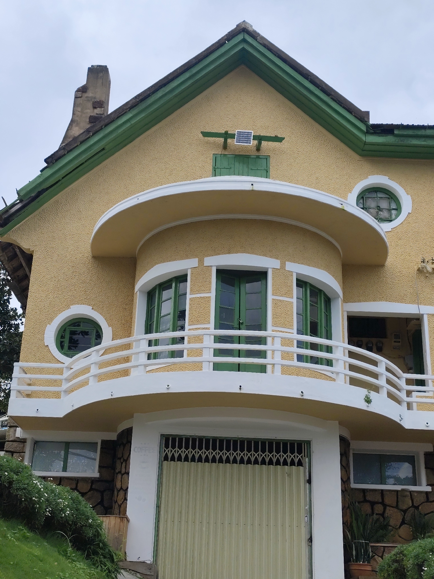 The image shows a close-up, exterior view of a yellow-colored house with green trim and accents, typical of the French colonial architectural style found in Da Lat, Vietnam. The house features a prominent curved balcony with a white railing, supported by a yellow rounded structure. Several windows are visible, including two large green-framed windows on the balcony, a smaller circular window above and to the left, and a larger circular window on the upper level to the right. The house has a textured stucco exterior and a dark tiled roof, parts of which are damaged. A disused brick chimney is visible on the left side of the roof. A small solar panel is mounted on the wall beneath a green-shuttered window on the upper level. The ground floor has a roll-up metal gate and a stone-clad wall with a small window. Lush green vegetation, including grass and flowering bushes, surrounds the lower part of the house, indicating a natural, possibly hilly, environment consistent with Da Lat's landscape. The sky is overcast, suggesting a cloudy day. The image appears to be taken from a low angle, looking up at the house.