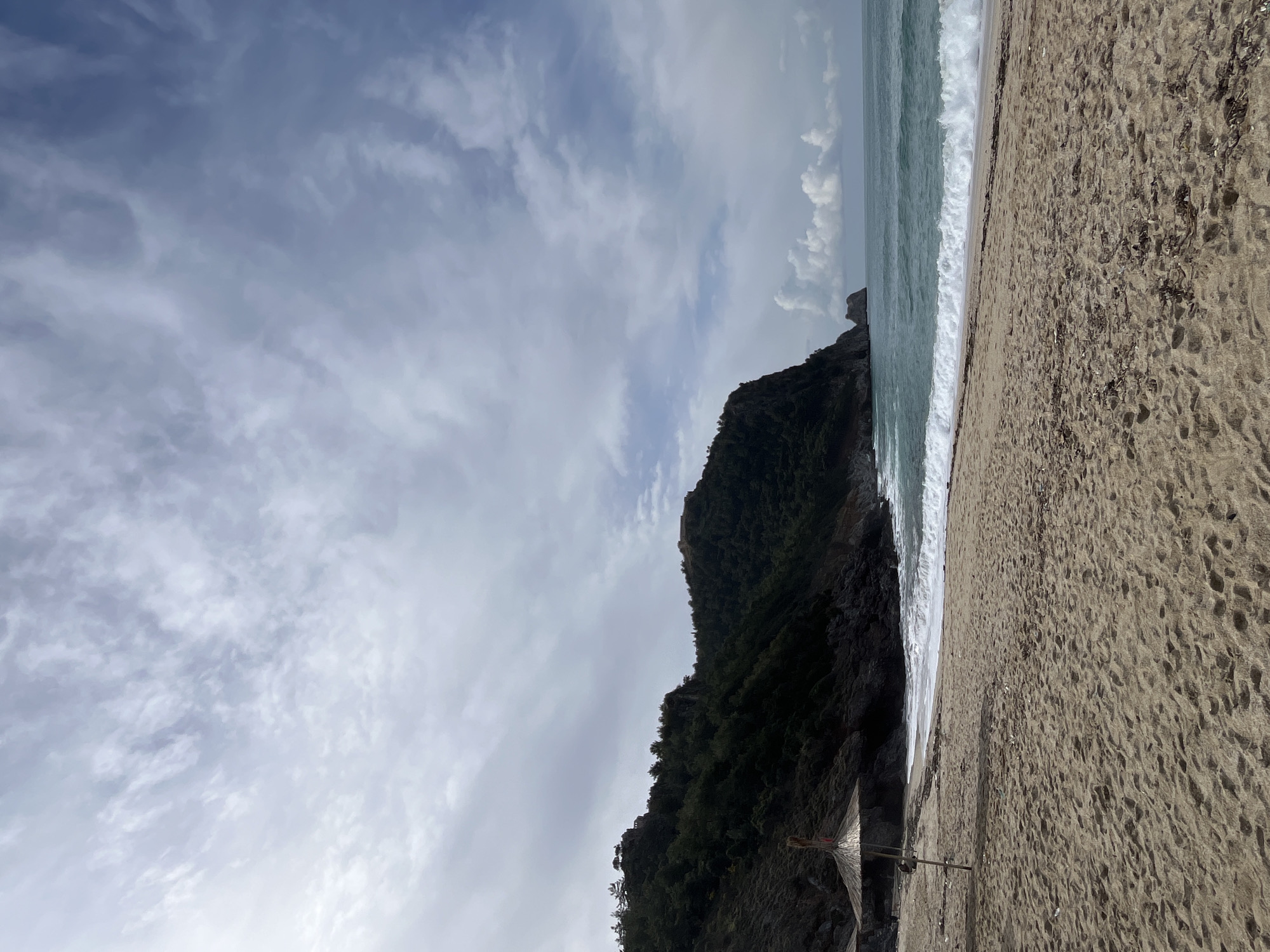 The image depicts a beach scene in Alanya, Turkey. The sky is overcast with cloudy formations. A rugged, steep, green, tree-covered hillside borders the sandy beach. Waves crash along the shoreline where the beach meets the turquoise sea. The sandy beach stretches into the foreground. There are no people immediately visible in the frame. A small, partially obscured structure, possibly part of a beach facility, is visible at the base of the cliff.