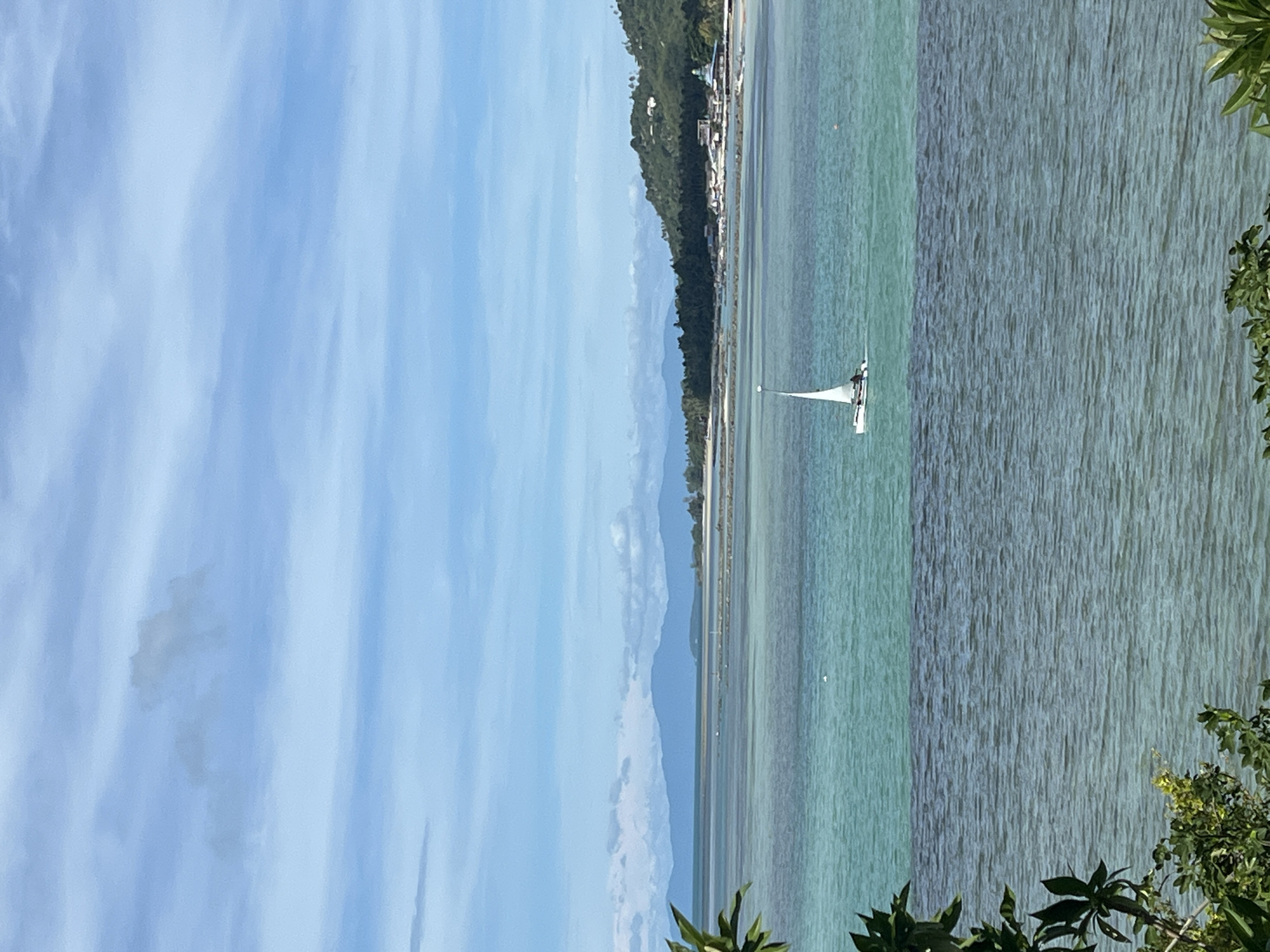 The image depicts a tranquil coastal scene in Baan Hua Thanon, Thailand. In the foreground, lush green foliage partially obscures the view of a calm, deep blue body of water. Beyond this, a strip of lighter, turquoise water is visible, suggesting shallows or a change in depth. In the mid-ground, a small white sailboat with a single mast is gently navigating the turquoise water. The horizon is marked by a distant, verdant coastline, with a range of muted blue mountains forming a backdrop. Scattered along the coastline are hints of buildings and trees. The sky above is a pale blue, adorned with wispy white clouds that stretch across the frame, indicating a pleasant day.