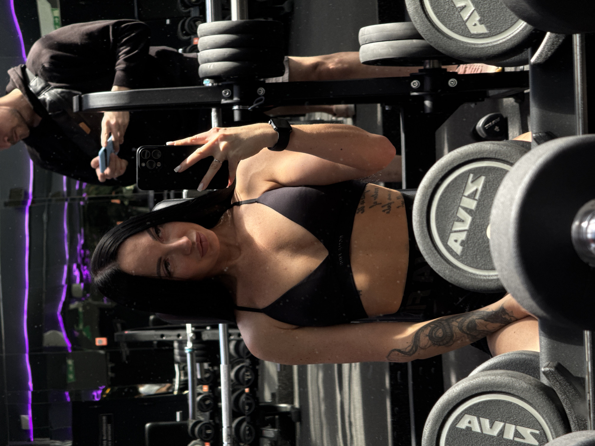 A mirror selfie captured in a gym in Kazan, Russia, features a woman in the foreground and a man reflected in the background. The image is oriented sideways, presenting the subjects and gym equipment horizontally.
The central subject is a woman with long dark hair, wearing a black sports bra. She looks directly at the camera with a neutral expression, holding a black smartphone (likely an iPhone) to take the picture. A smartwatch is visible on her right wrist, and a ring adorns her left ring finger. She has a distinctive, intricate tattoo on her left forearm, resembling a serpentine or dragon-like design, and faint text tattooed on her right ribcage.
In the background, a man wearing a dark hoodie is partially visible, also reflected in the mirror. He appears to be engaged with gym equipment, holding a light blue object in his hand.
The setting is a well-equipped fitness center, evident from the various weight plates, dumbbells, and racks. Several weight plates clearly display the brand "AVIS." The gym also features purple LED strip lighting, adding a modern touch to the ambiance. What appears to be natural light from an unseen source illuminates parts of the scene, suggesting it is daytime. The overall activity is a casual gym visit, with one person taking a selfie and another working out.
