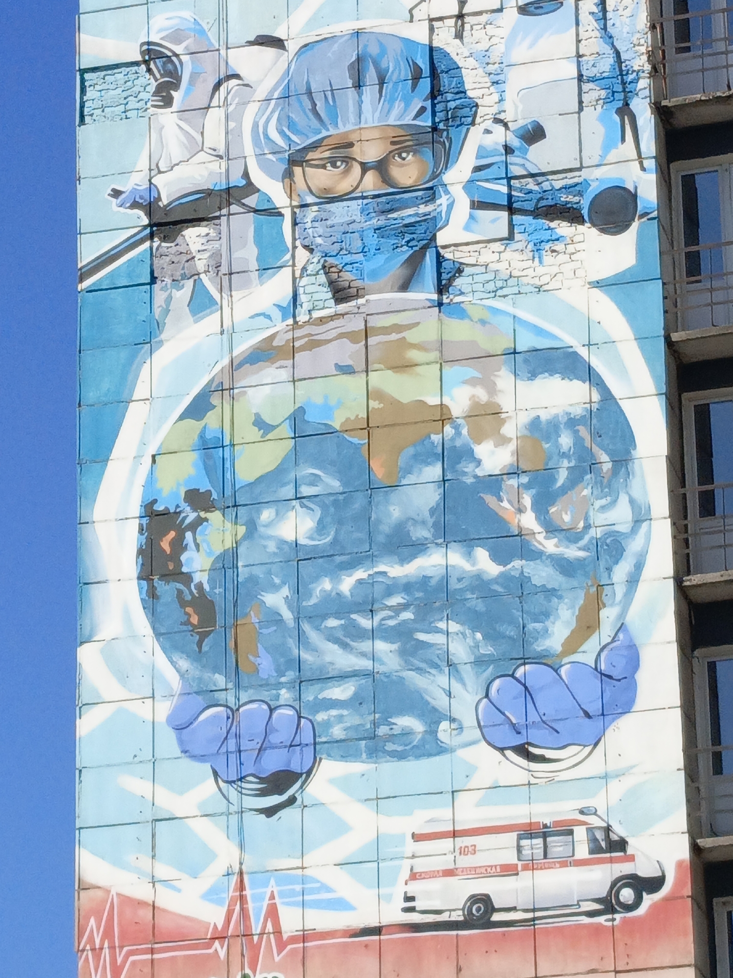 The image shows a mural on the side of a building in Perm, Russia. The mural depicts medical personnel in protective gear, a doctor in a surgical mask and cap, holding a globe. An ambulance is at the bottom, with an electrocardiogram graphic. The sky above is blue, and a portion of the building's windows is visible.
