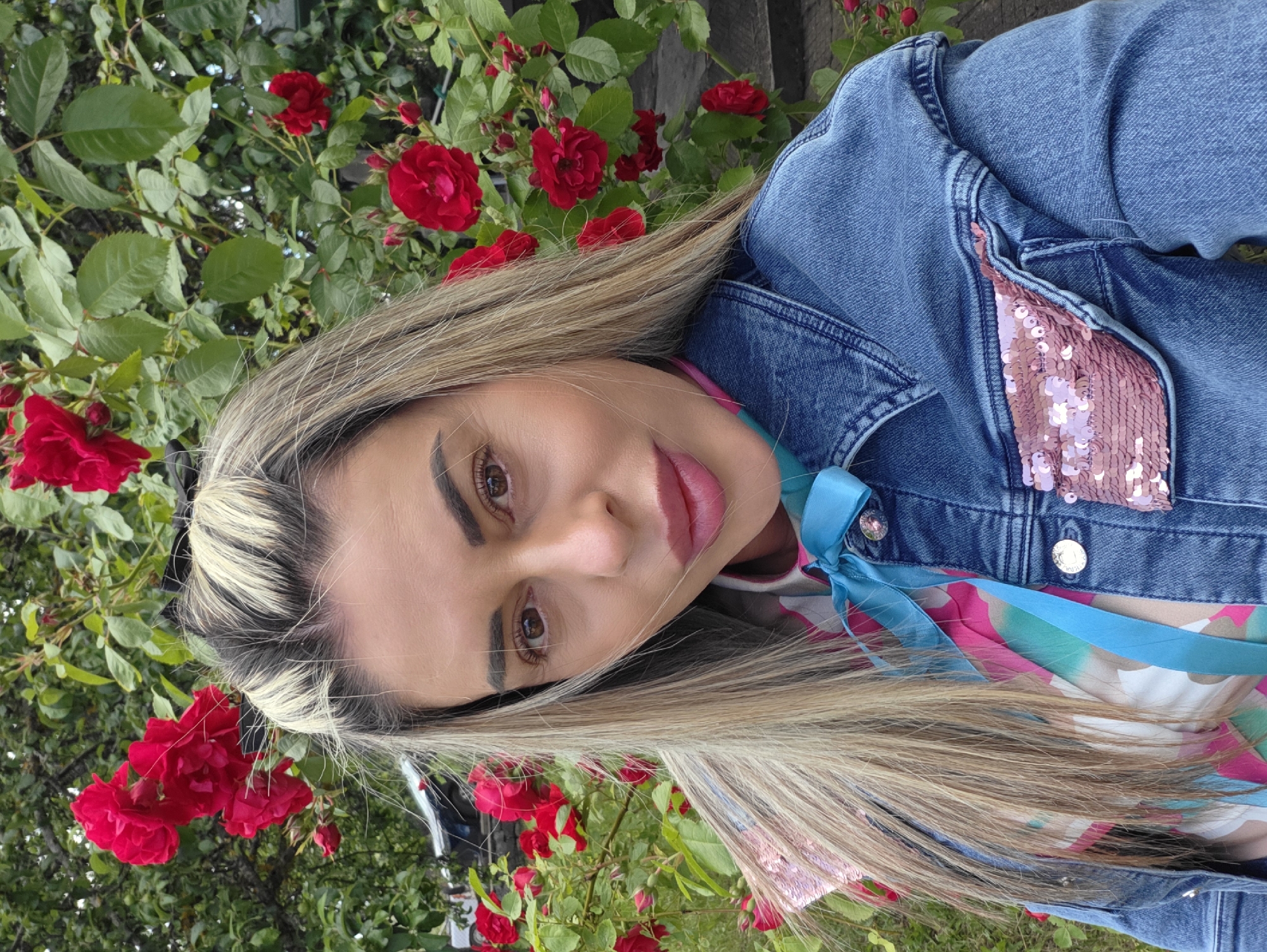 A medium shot photograph features a woman standing outdoors in front of a rose bush. The woman is positioned in the center of the frame and is looking towards the camera. She has long, blonde hair, some of which is pulled back. Her eyes are brown, and she is wearing makeup. She is dressed in a denim jacket with pink sequin embellishments on the pockets, and a colorful patterned shirt with a blue ribbon tied in a bow around her neck. The rose bush behind her is in bloom with vibrant red roses and green leaves. The background is slightly blurred, but it appears to be a natural setting with hints of greenery and possibly a pathway or ground covered with stones. The overall lighting suggests it is daytime. There is no indication of the specific location in the city of Ravnogor, Bulgaria, within the image itself.