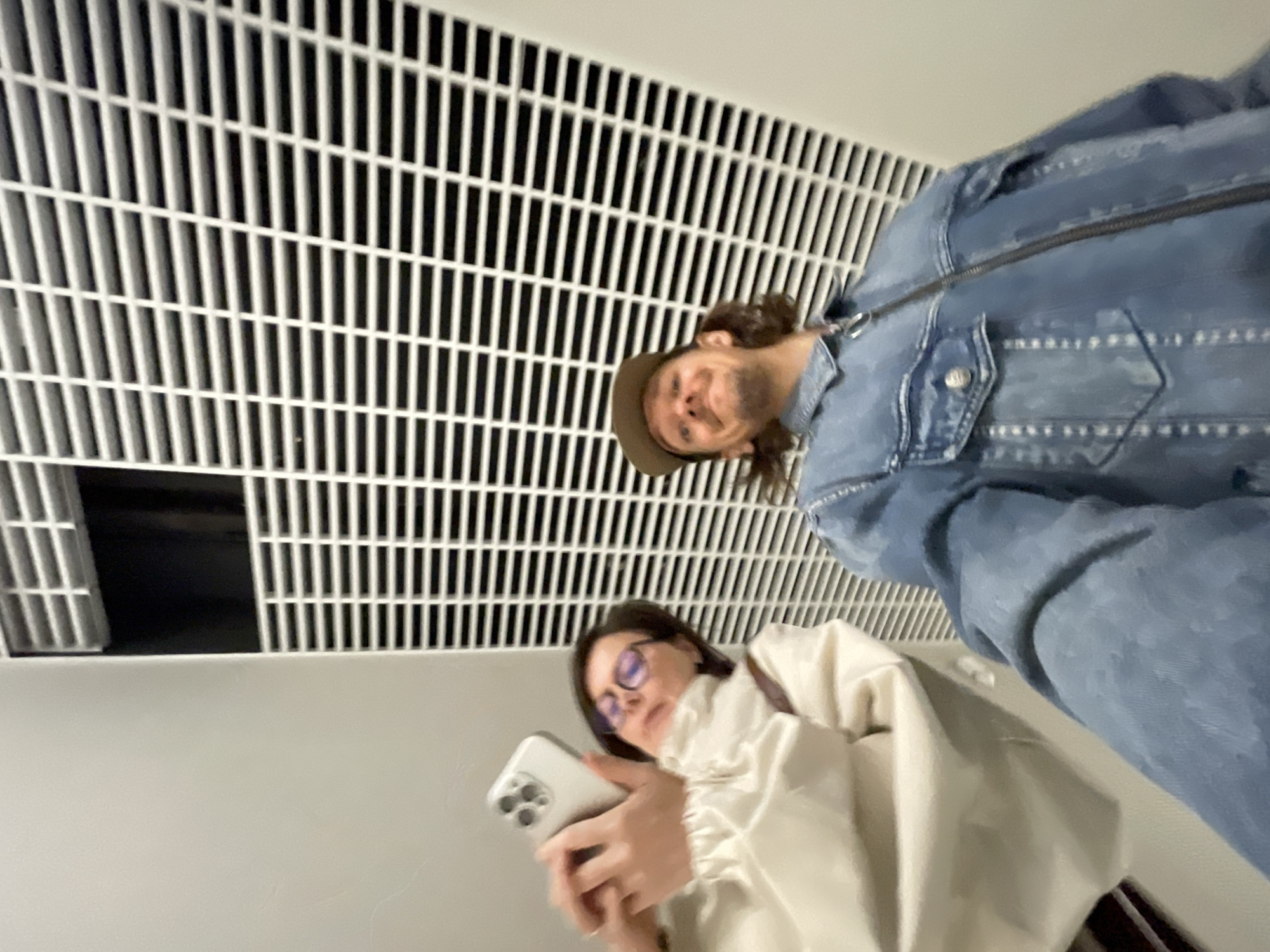 The image appears to be taken from a low angle, looking upwards. A white, grated ceiling or vent system dominates the upper portion of the frame. Two individuals are present. One person, wearing a denim jacket and a hat, is positioned to the right and slightly above the other. The second person, wearing glasses and a light-colored jacket, is looking at a mobile phone they are holding. The background is primarily white. There is no information suggesting the location is Izhevsk, Russia.