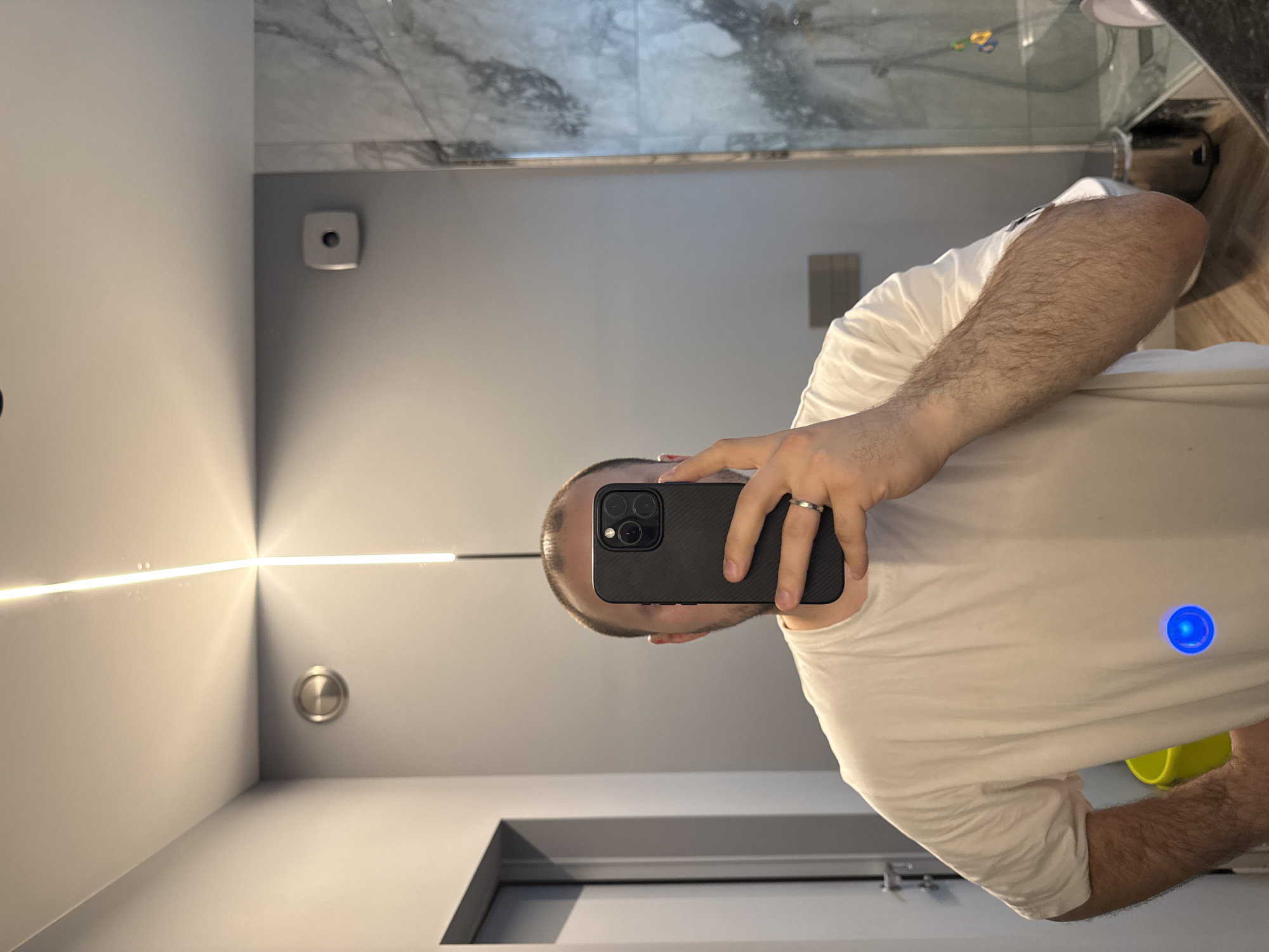A person stands in a bathroom taking a selfie in the mirror. The bathroom has a white marble shower, gray walls, a vanity, and a linear LED light. The person is wearing a white T-shirt and holding a dark colored cell phone. It is not possible to determine the location of the bathroom as being in the city of Krasnoyarsk, Russia.