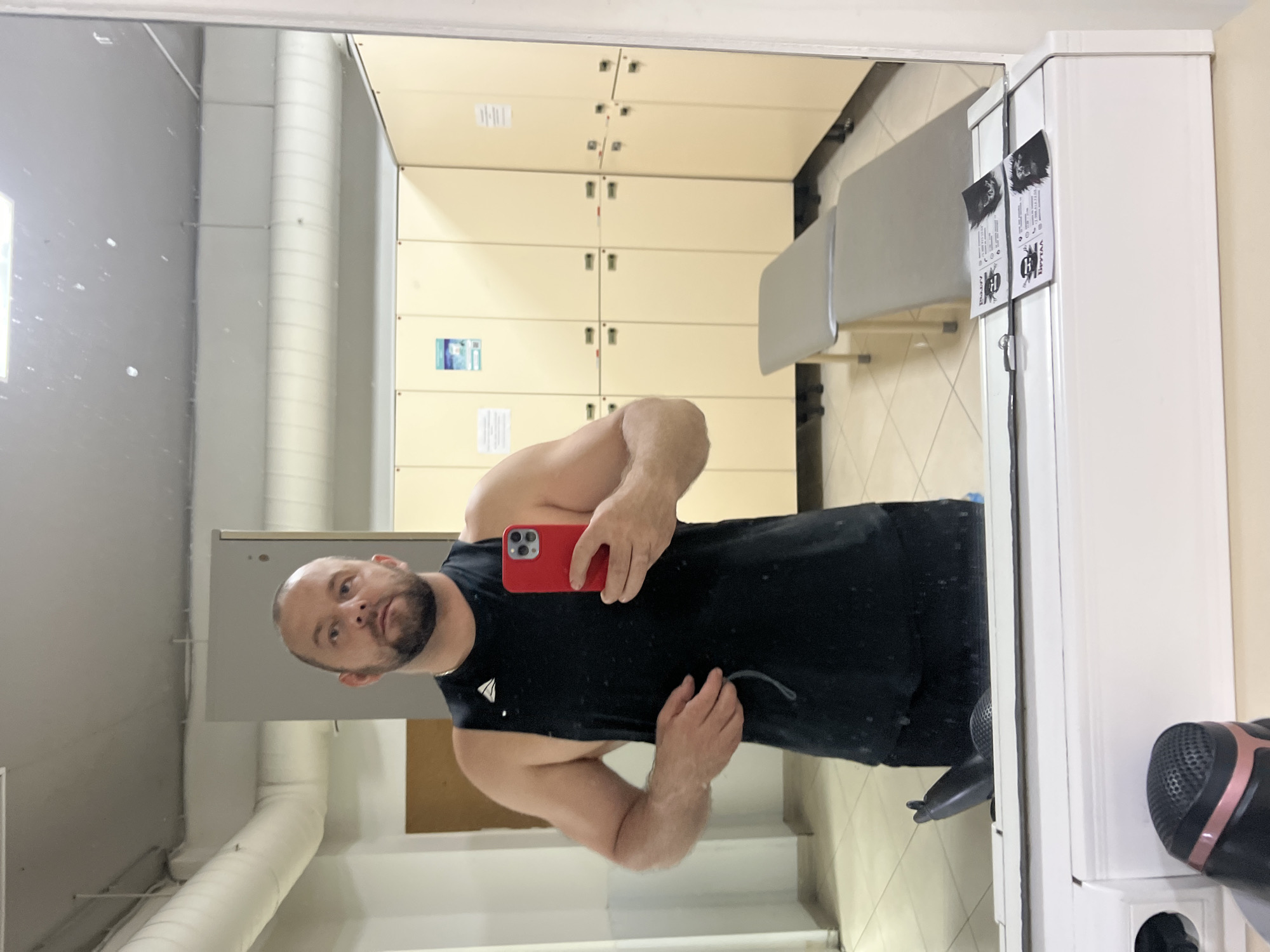 The image is a mirror selfie taken in what appears to be a changing room or locker room, likely within a gym or fitness center in Sochi, Russia. A man, seen from the chest up, is the primary subject. He has a short beard and a bald head, and is wearing a black sleeveless top with a small white, stylized logo on the left chest, resembling a sailboat. He holds a red smartphone to capture the reflection.
The background, seen through the mirror, features rows of light beige or cream-colored lockers with silver handles. Two simple, light gray benches are positioned in front of some lockers on the right side of the reflection. Along the left wall, a large white cylindrical ventilation duct is visible. The floor is covered with light-colored tiles.
On the right side of the mirror frame, two identical white tags are hung. The most prominent text in Cyrillic on these tags is "БУЗЫГ," followed by smaller text "БИЗЫМ" and graphic elements, including what appears to be a bee. A black and pink hairdryer is partially visible on a counter or shelf to the immediate right of the mirror. The setting is indoors and well-lit.