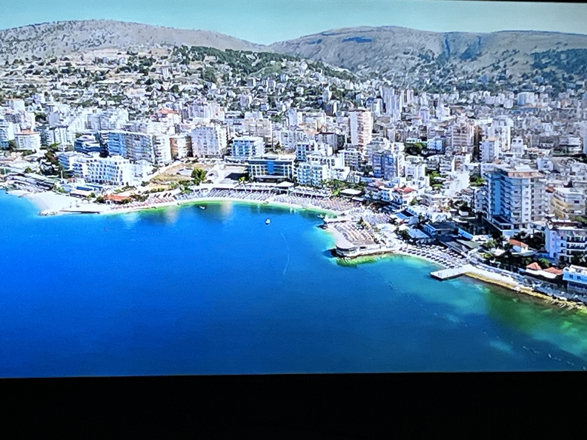 This is an aerial, high-angle, wide shot of a coastal city with mountains in the background. The city is densely packed with buildings, mostly white and light-colored, with some featuring blue accents. A significant portion of the foreground is occupied by a large body of clear blue water, transitioning to a greenish hue closer to the shore, indicating shallow areas or a rocky bottom. Along the coastline, there is a beach lined with numerous rows of white umbrellas and lounge chairs, suggesting a resort or popular tourist area. Several small boats are visible on the water. The city appears to be built on a slope leading up to the mountains, which are covered in sparse vegetation. There are no visible people or specific landmarks that would identify the location as Kharkiv, Ukraine.