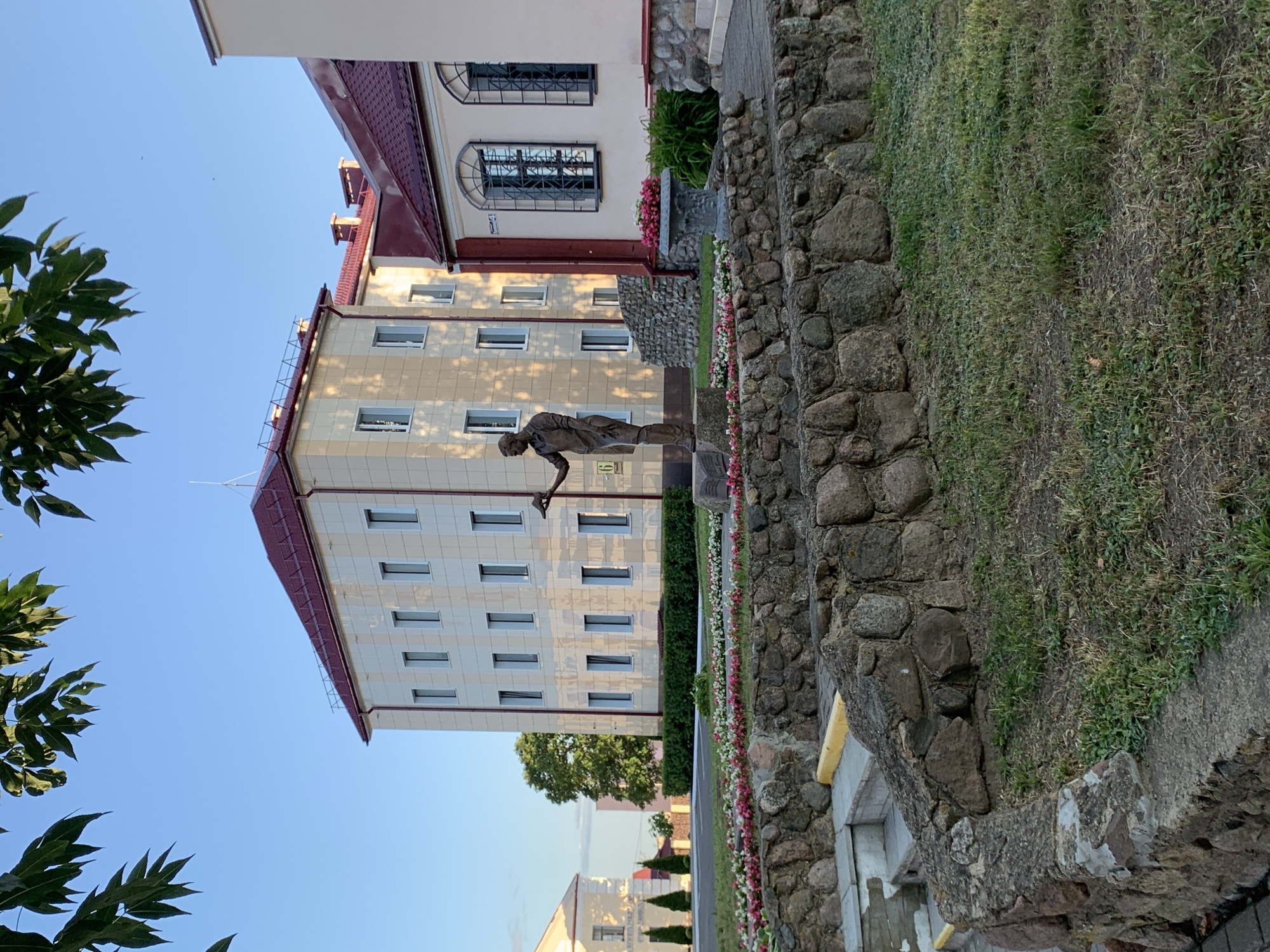 The image depicts a scene in Kapyĺ, Belarus, featuring a bronze statue of a man standing in front of two buildings. The statue, placed on a stone pedestal, shows the man holding a palette and brush, suggesting he is an artist. Behind him, a light-colored, multi-story building with a red roof dominates the background. To the right, another building with a red roof and arched windows with decorative grilles is visible. The foreground is characterized by a tiered stone wall bordering a grassy area with a flowerbed. The sky is clear and blue, with green leaves from an overhead tree partially obscuring the top of the image. The overall ambiance is serene and suggests an outdoor public space.