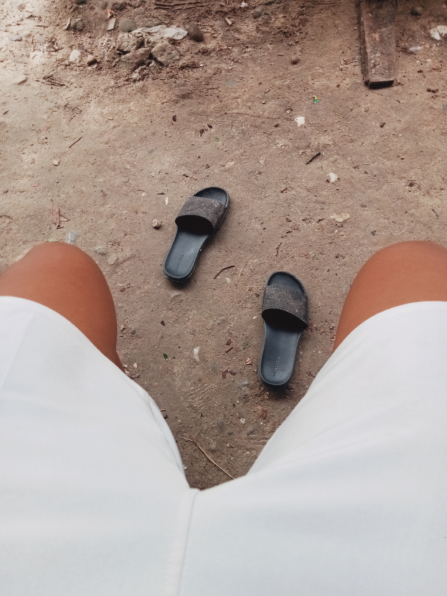 A first-person perspective shot shows a pair of black slides with textured straps lying on dirt ground. The slides are placed to the left and right of a person's legs, which are clad in white shorts. The ground is composed of brown soil, small rocks, and scattered debris, suggesting an outdoor environment, possibly in a park or unpaved area. The lighting suggests it's daytime. The image does not provide any specific identifiable landmarks or features that would indicate a location in Pamesi, Indonesia.