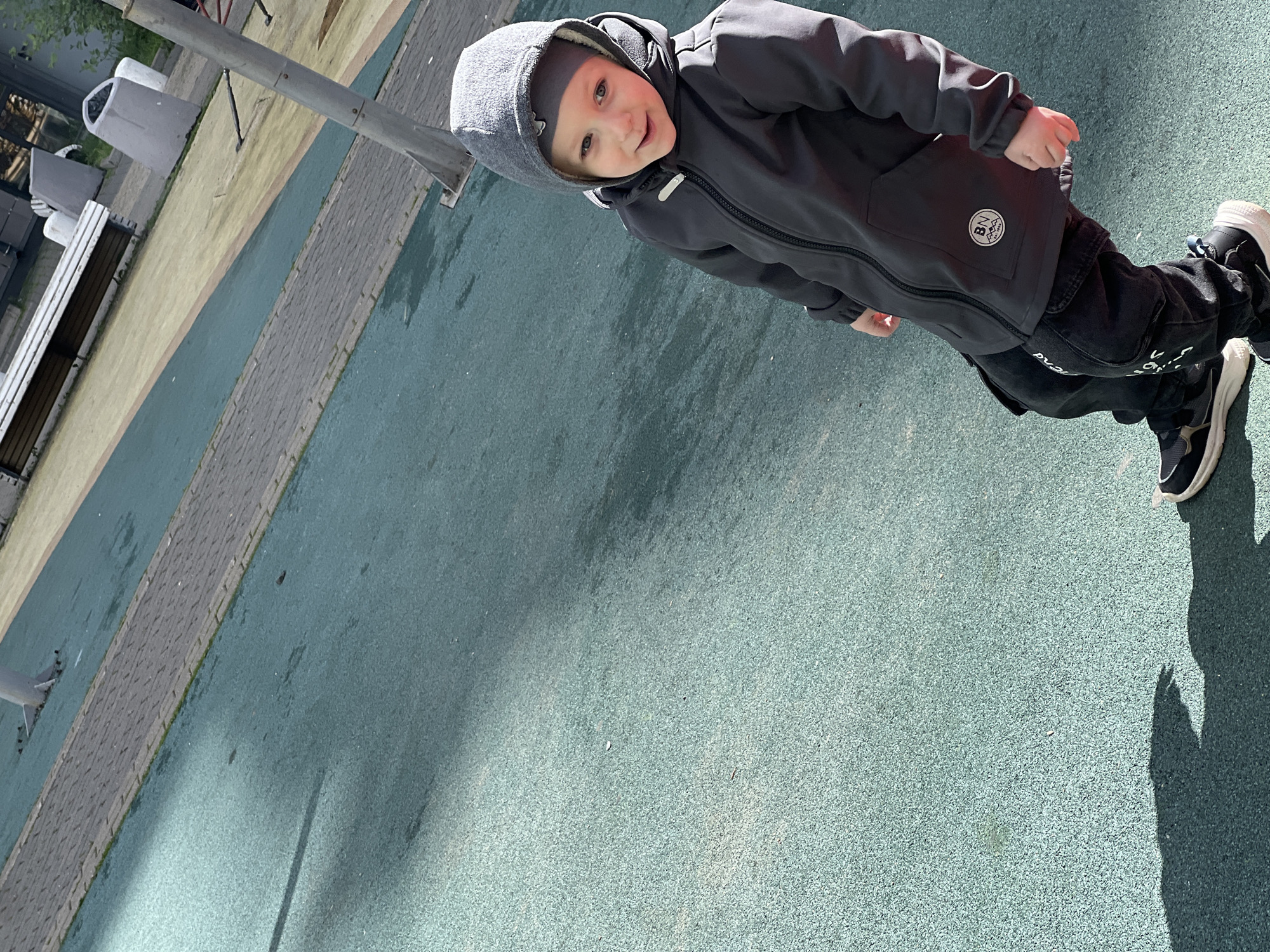 A young child wearing a gray hooded jacket and dark jeans stands on a green surface, possibly a playground or park area. The location appears to be in an urban setting, likely in the city of Mytishchi, Russia. A park bench and a lamppost are visible in the background. The child is looking toward the camera and appears to be smiling.