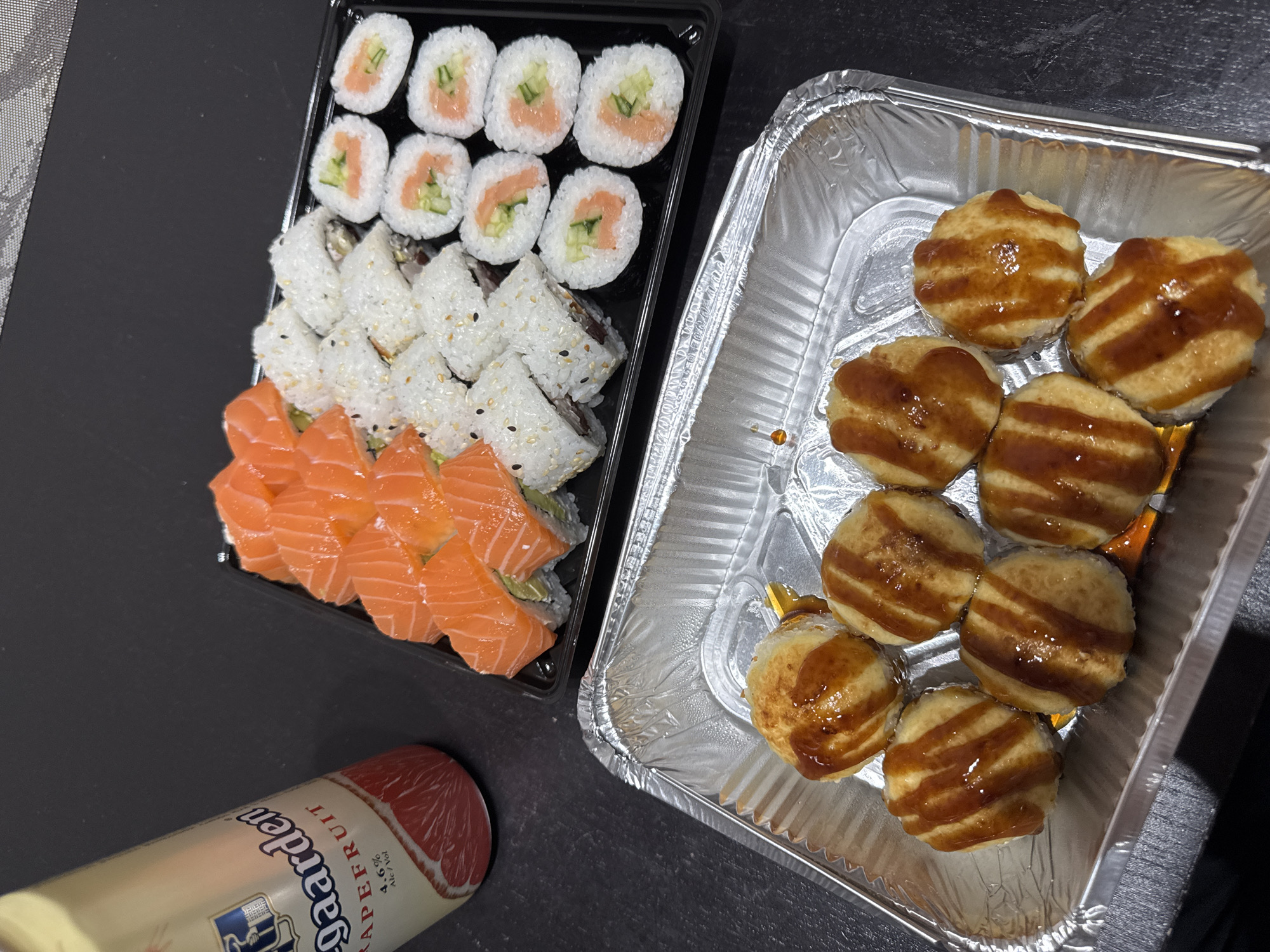 An overhead shot captures a takeaway meal arranged on a dark, textured surface, likely a table. The scene features a black plastic tray filled with various types of sushi and a silver aluminum foil container holding baked items.
The sushi tray contains three distinct rows. The top row features eight pieces of hosomaki, cylindrical sushi rolls with seaweed on the outside, filled with salmon and cucumber. The middle row displays eight uramaki (inside-out rolls) coated in white rice and sprinkled with black sesame seeds. The bottom row consists of eight pieces of salmon-topped rolls, where slices of fresh, vibrant orange salmon are draped over what appear to be rice rolls, possibly with an avocado filling visible beneath.
To the right, an oval-shaped aluminum foil container holds eight individual, round, golden-brown items. These items, possibly small baked pastries, pies, or a unique type of cooked sushi, are each generously drizzled with a glossy, dark brown sauce applied in striped patterns.
In the bottom left corner, a cream-colored can of "Hoegaarden GRAPEFRUIT" beer is partially visible, oriented horizontally. The text "4.6% Alc./Vol." is also clearly legible on the can.
The setting appears to be an indoor, casual dining environment, possibly at home. No people are visible, and no specific time of day or external location cues are present in the image itself.