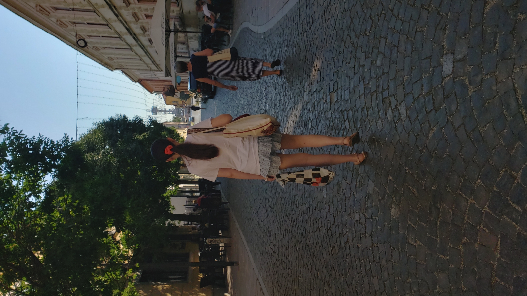 The image depicts a cobblestone street in the city of Prešov, Slovakia. In the foreground, a young person with a ponytail walks away from the camera, wearing a white t-shirt, a patterned skirt, and sandals. They are carrying a checkered reusable bag in their left hand and a tote bag over their right shoulder. Behind them, an adult wearing a striped dress also walks away from the camera. The street is lined with buildings on the right, some of which appear to be shops or cafes with outdoor seating. Trees with green foliage are visible on the left side of the street. In the distance, a tall tower is visible against a clear blue sky. The street is illuminated by sunlight, casting shadows on the cobblestones.