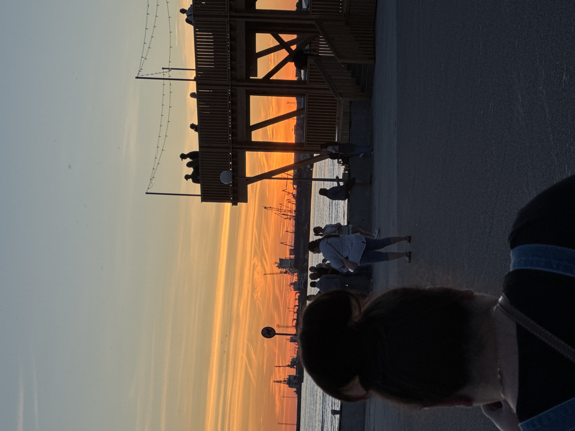The image captures a sunset scene at a harbor in Tallinn, Estonia. Silhouetted against the vibrant orange and yellow sky are numerous cranes and ships, suggesting an active port. In the foreground, a wooden, multi-level structure with railings and stairs is prominent, serving as a viewing platform. Several people are visible, some standing on the platform, others gathered near the water's edge, and one person is silhouetted in the very foreground with their back to the camera. The ground is paved with asphalt, and the sea is calm. String lights are visible along the top of the wooden structure.